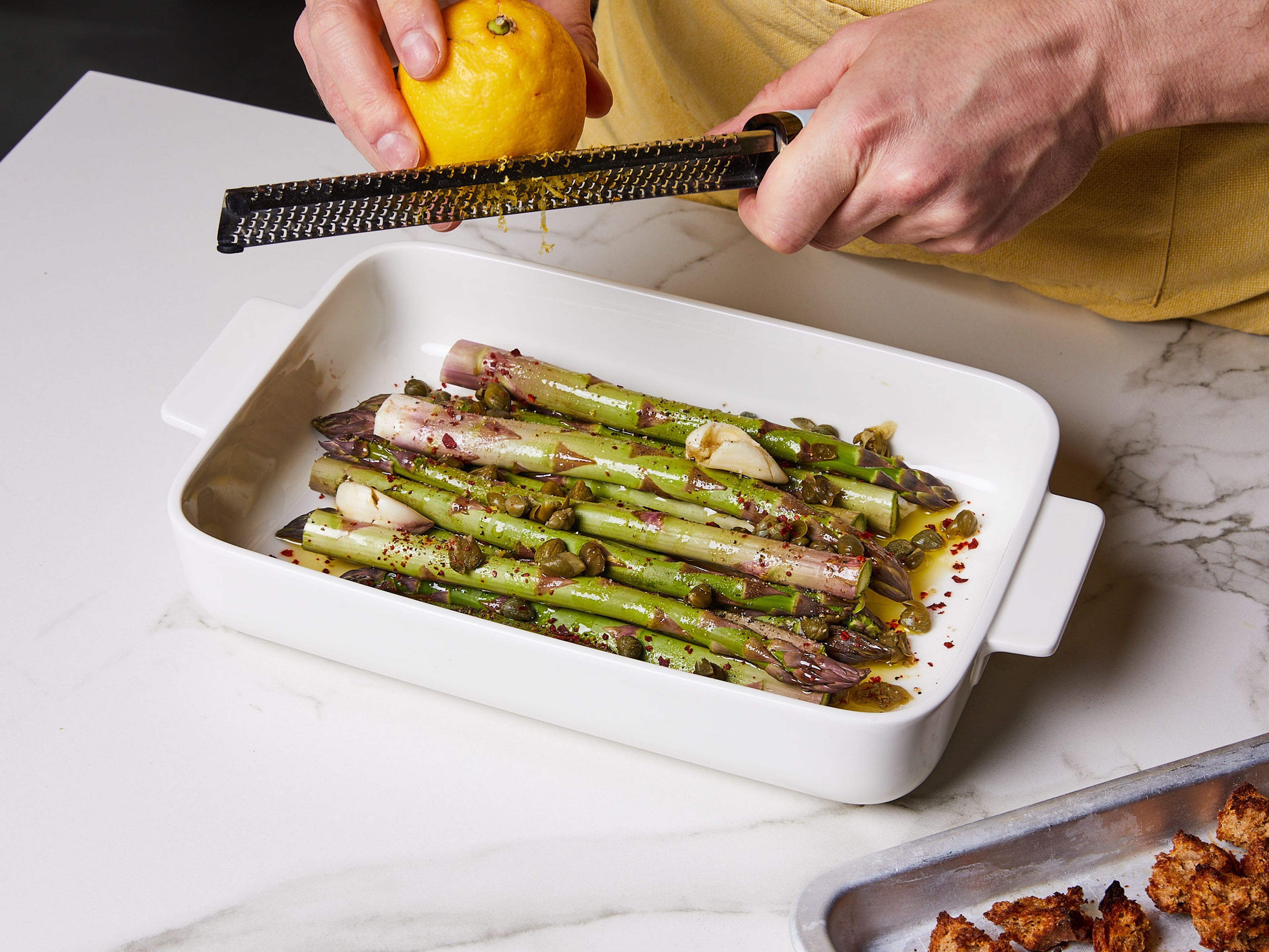 Mix olive oil, asparagus, capers, lemon zest, garlic, chili flakes, salt, and pepper in a baking dish. Place in the oven to roast for approx. 18–24 min. until the asparagus is lightly browned.