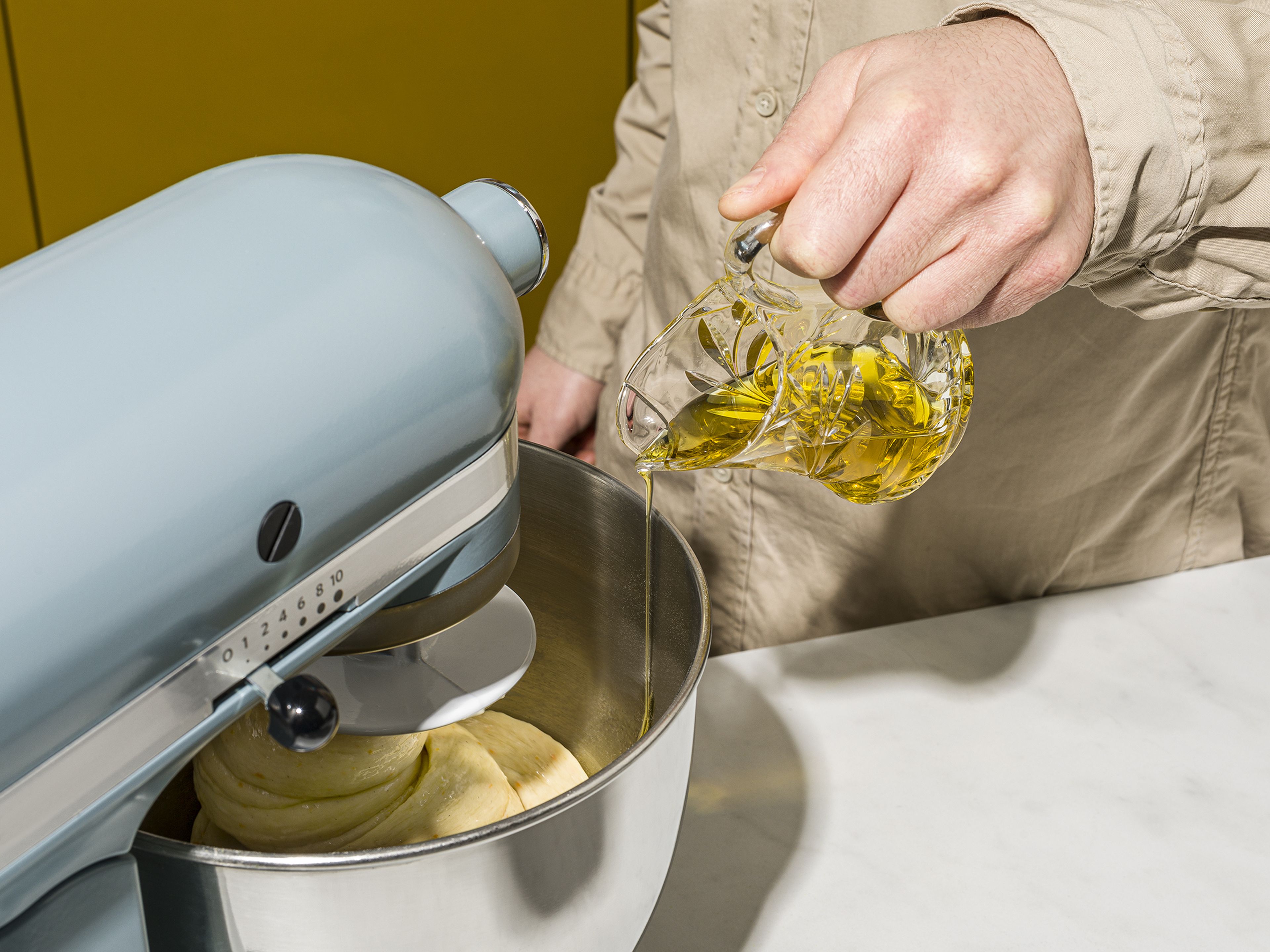 Für den Grundteig restliches Mehl mit Zucker und der restlichen Hefe, Honig, Milch, einem Teil der Eier, Orangenabrieb und Vanilleextrakt in einer Küchenmaschine verrühren, bis sich ein gleichmäßiger Teig bildet. Nun der Reihe nach den Vorteig, anschließend das Salz und zuletzt ein Viertel des Olivenöles hinzufügen. Sobald die Zutaten vollständig in den Teig eingearbeitet sind, kann das restliche Öl untergemischt werden. Teig für weitere 5 Min. bei hoher Geschwindigkeit in der Maschine kneten.