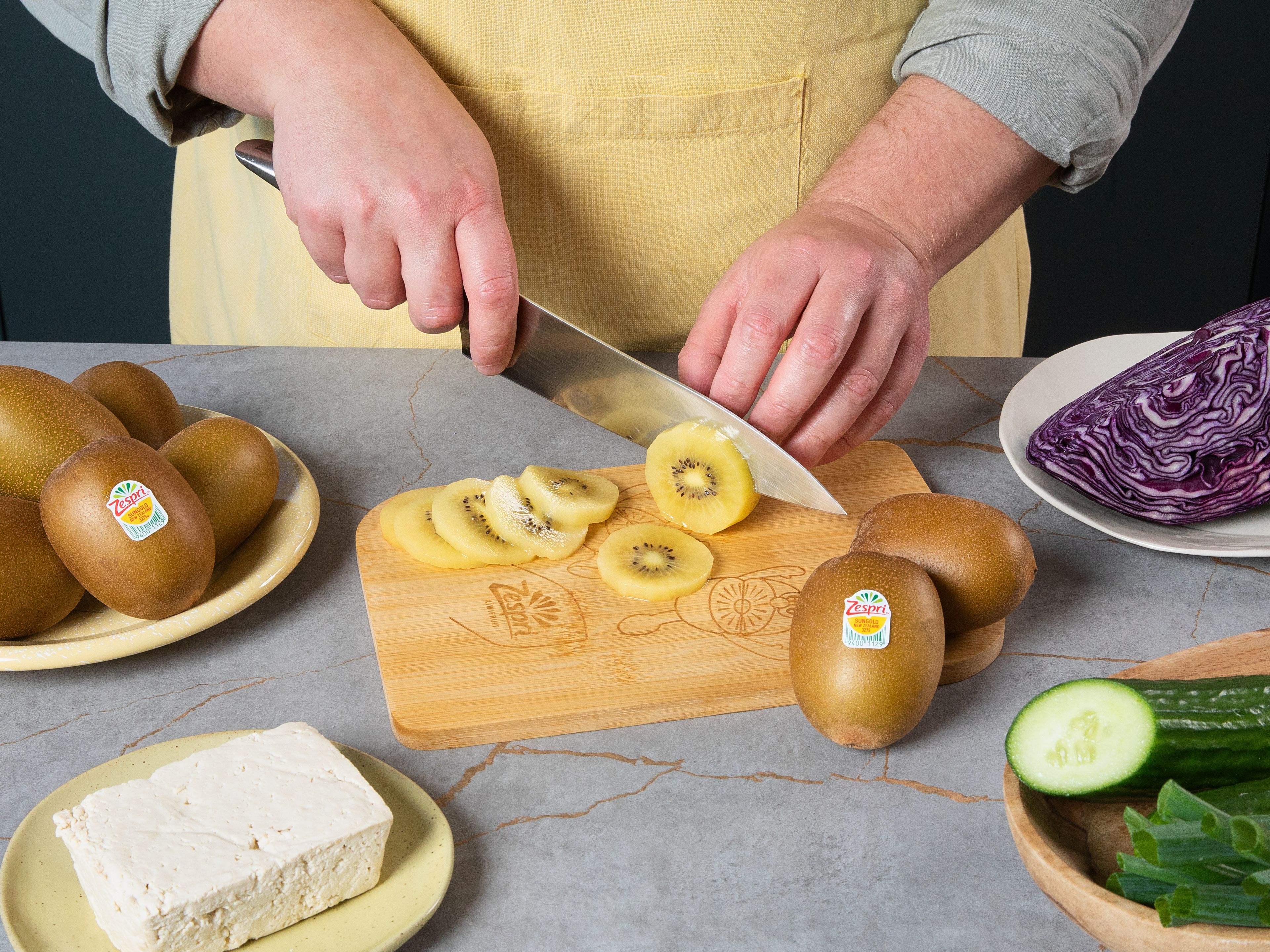 Tofu in ein Küchentuch wickeln und für ca. 10 Min. mit einem schweren Gegenstand beschweren. In der Zwischenzeit Zespri™ SunGold™ Kiwis schälen und in Scheiben schneiden. Gurke ebenfalls in dünne Scheiben schneiden und Frühlingszwiebeln in feine Ringe schneiden. Den Rotkohl fein hobeln. Den abgetropften Tofu  in ca. 2 cm große Würfel schneiden. Sobanudeln nach Packungsanweisung kochen. Edamame ca. 3 Min. vor Ende der Garzeit mit ins Nudelwasser geben. Abgießen, kalt abschrecken und gut abtropfen lassen.