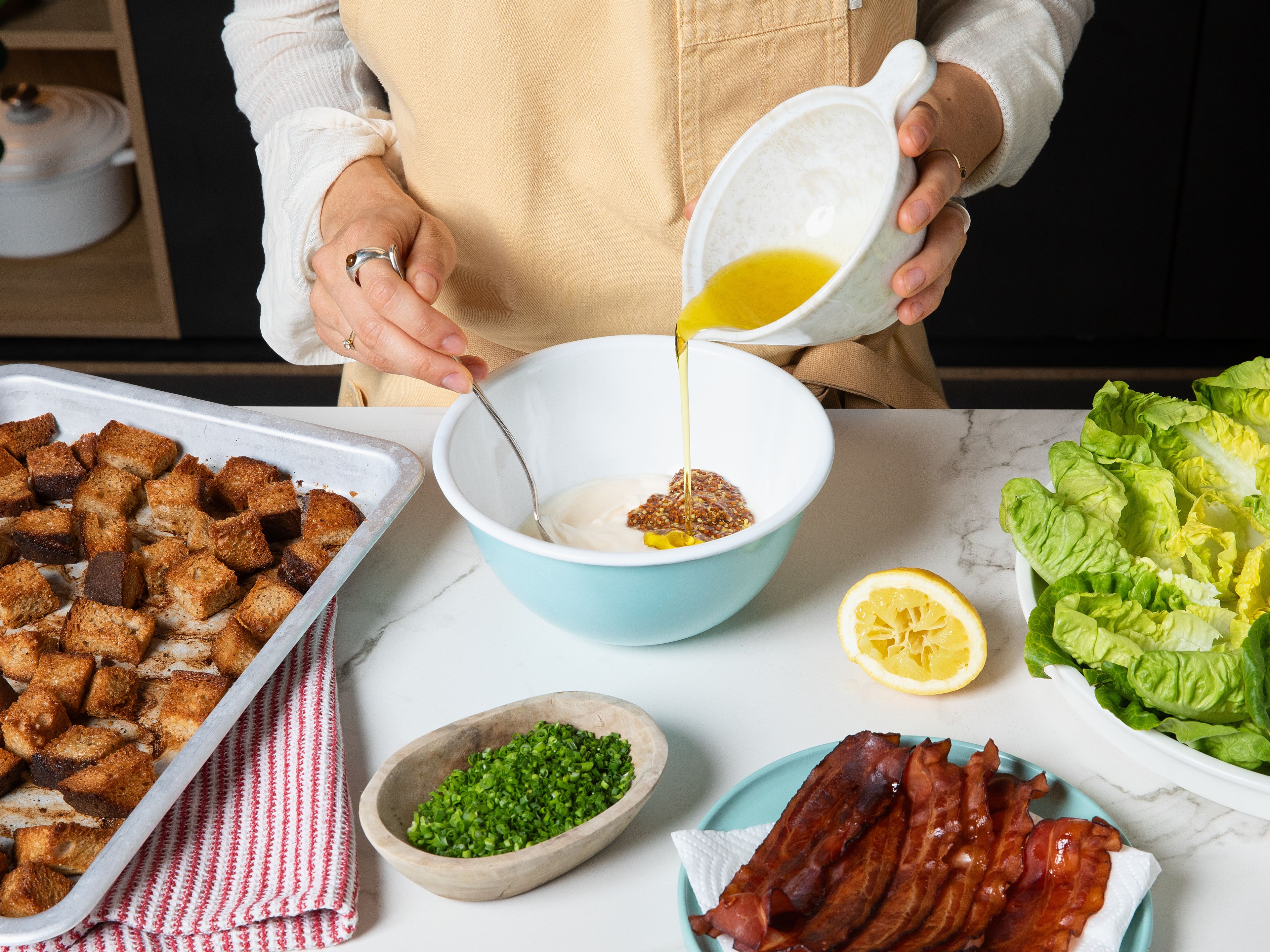 Für das Dressing saure Sahne mit Mayonnaise, Zitronensaft, Knoblauch und Senf in einer kleinen Schüssel verrühren. Olivenöl langsam einrühren, bis ein cremiges Dressing entsteht. Schnittlauch unterheben und mit Salz und Pfeffer abschmecken. Salatblätter auf einer Platte anrichten, Tomaten und Bacon in kleinen Stücken darauf verteilen, mit dem Dressing beträufeln und den Croûtons toppen.
