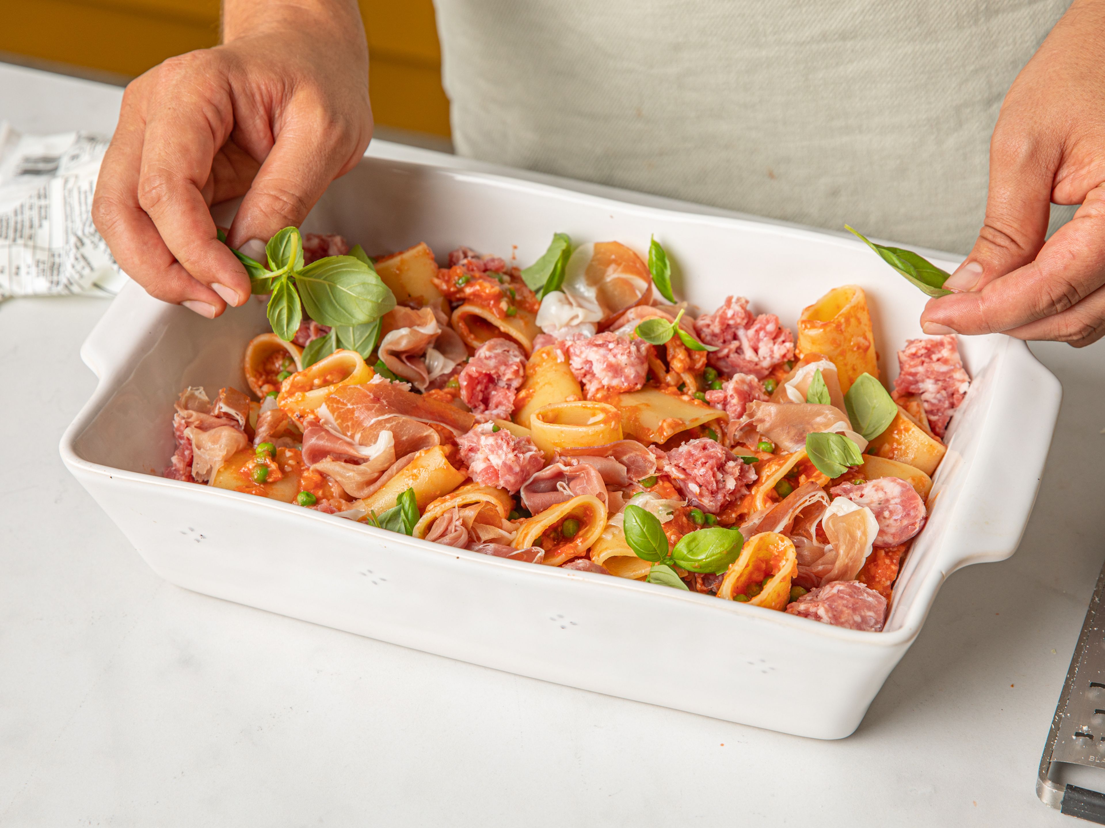 Den Backofen auf 200°C vorheizen. In einer großen Schüssel die Nudeln mit 2/3 des geriebenen Parmesans, den Erbsen und 2/3 der Tomatensoße vermischen. Dann in eine Auflaufform geben. Den Wurstdarm von der italienischen Wurst entfernen. Mundgerechte Wurststücke, Eier, Basilikum, Mozzarella und Prosciutto di Parma über die Nudeln streuen. Die verbliebene Tomatensoße und den restlichen Parmesankäse darüber geben.