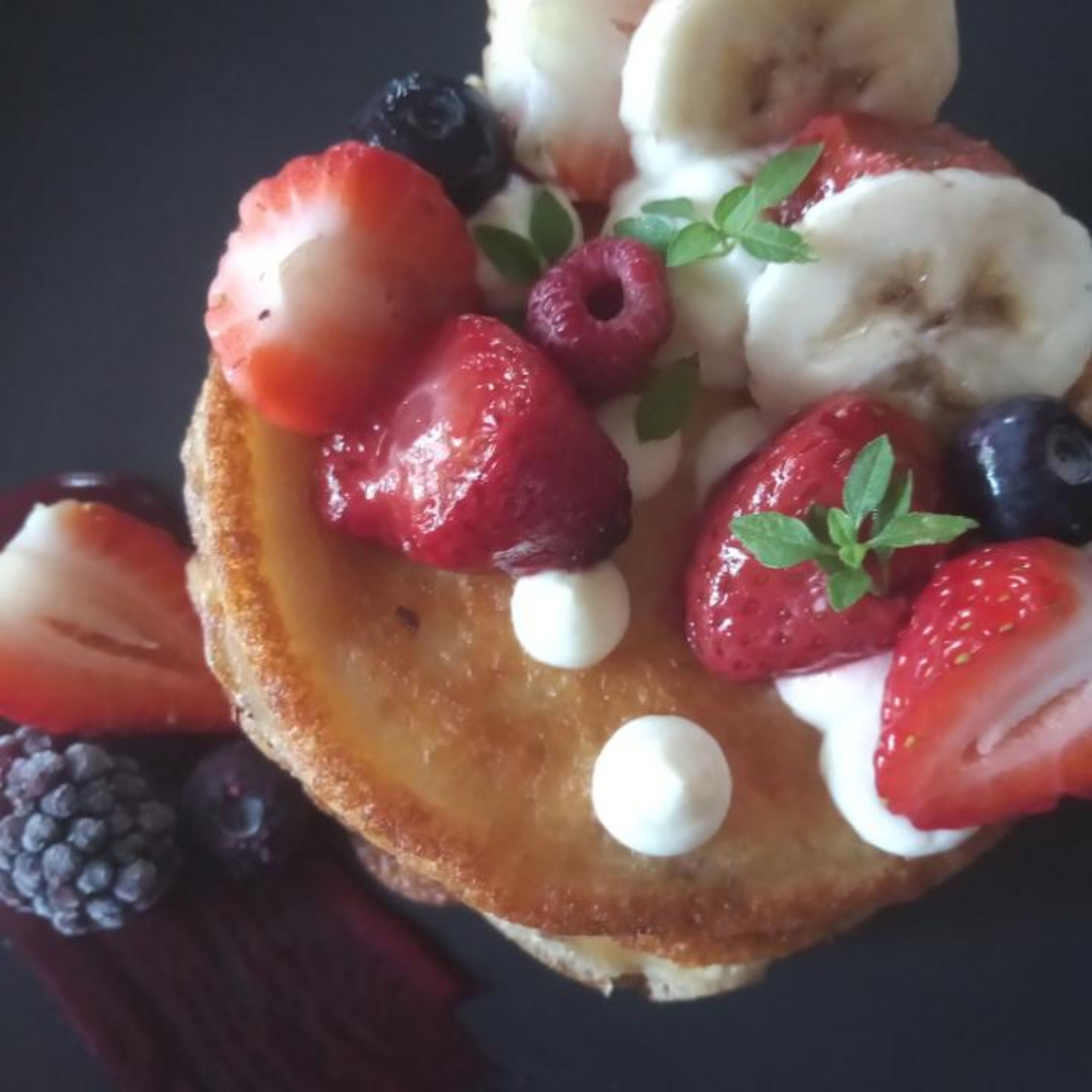 Plate up the dish. Start by stacking pancakes on a black plate, with a thin layer of whipped ricotta between each pancake. Pipe on whipped ricotta, add roasted strawberries and fresh fruit. Top with micro herbs and add berry puree.