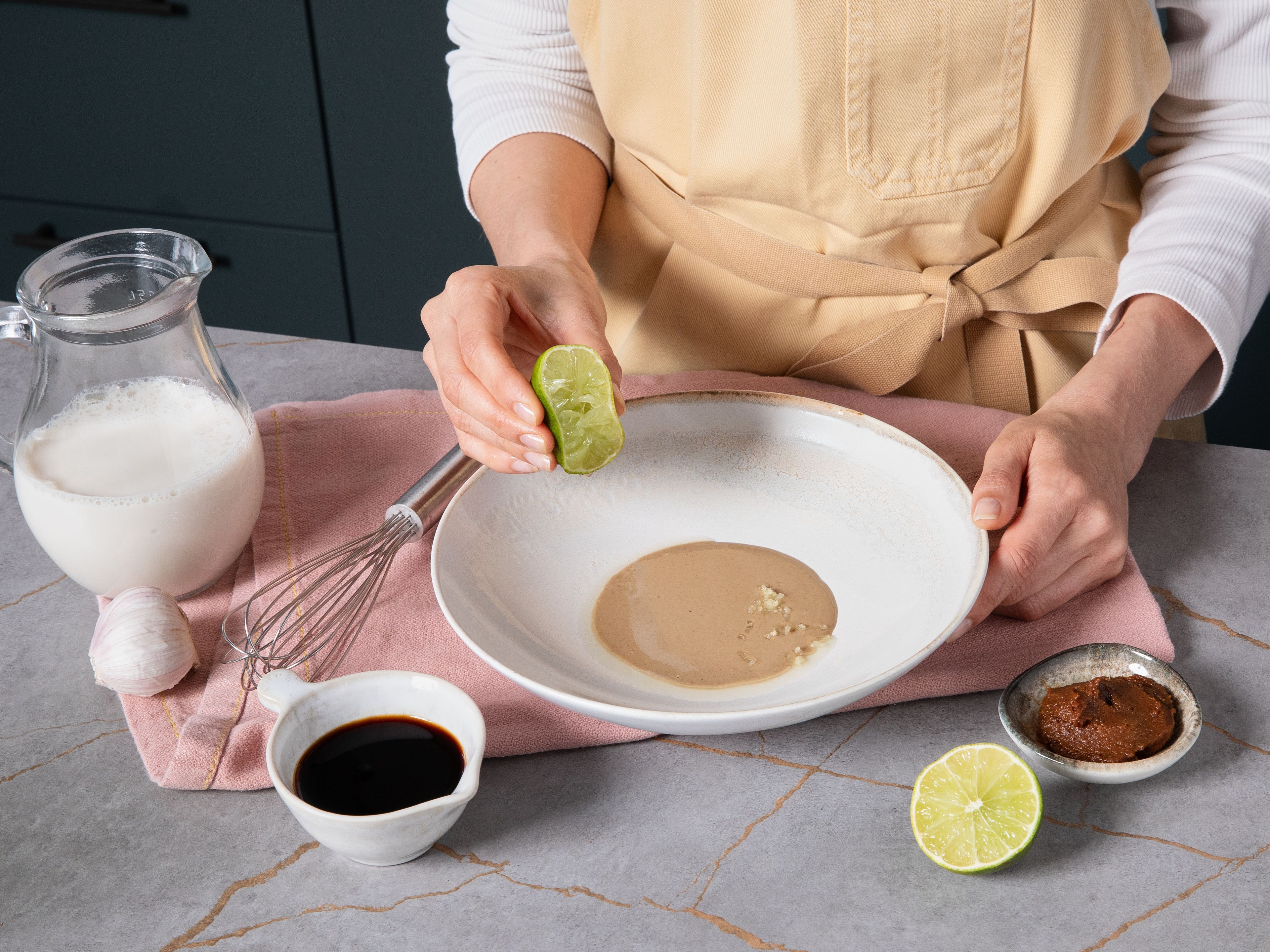Währenddessen die Tahini-Brühe mixen. Dafür direkt im Servierteller jeweils die Hälfte Knoblauch reiben und mit Tahini, Misopaste, Sojasauce, Limettensaft und Sojamilch verrühren, bis die Brühe cremig ist. Mit Salz und Zucker abschmecken.