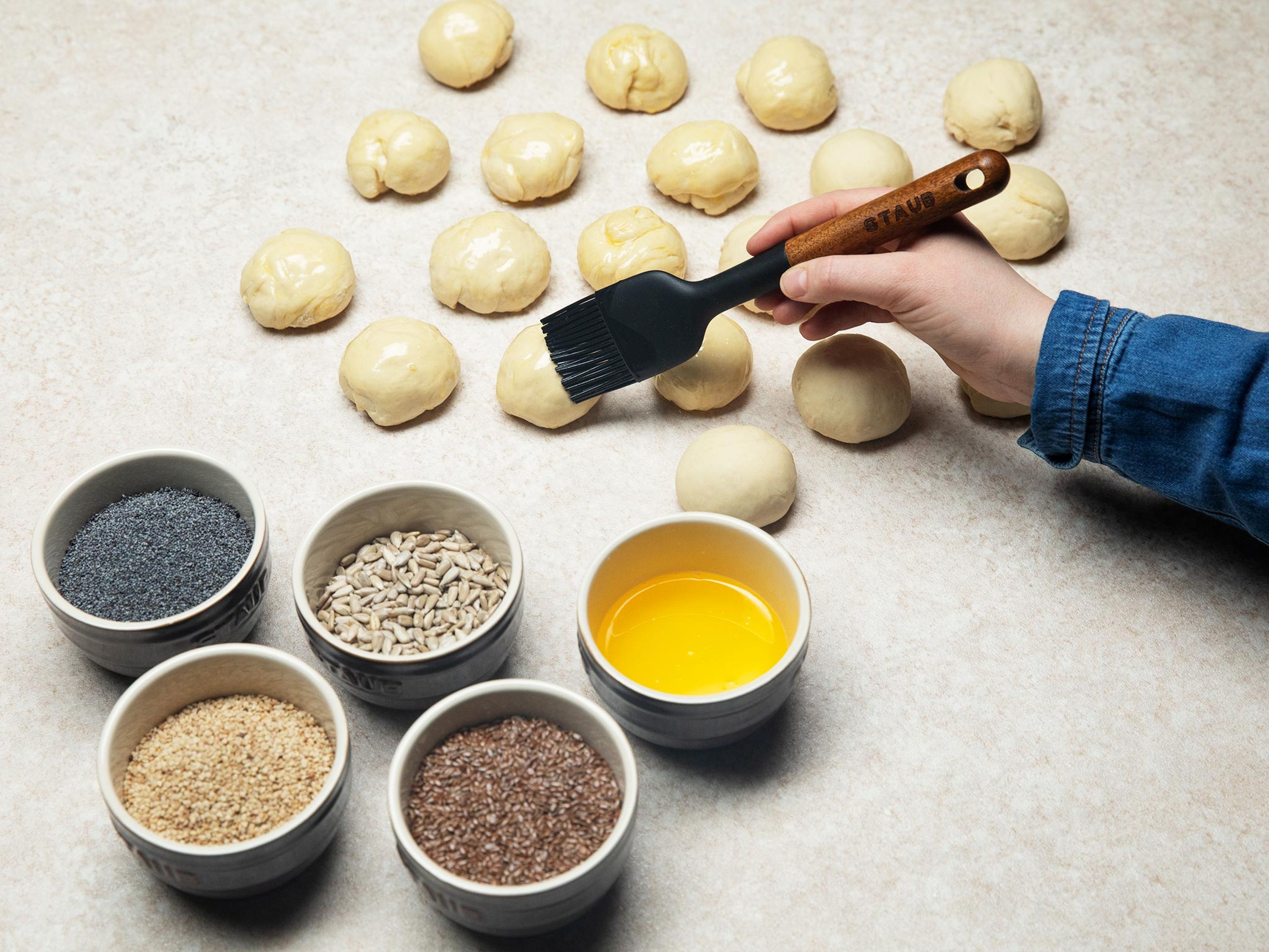 In the meantime, grease the bottom and sides of an oven-proof pan. After the dough has doubled in size, divide it into even pieces. Then brush melted butter on top and dip in either sesame seeds, poppy seeds, flaxseeds, or sunflower seeds—alternating the seeds as you brush and dip.