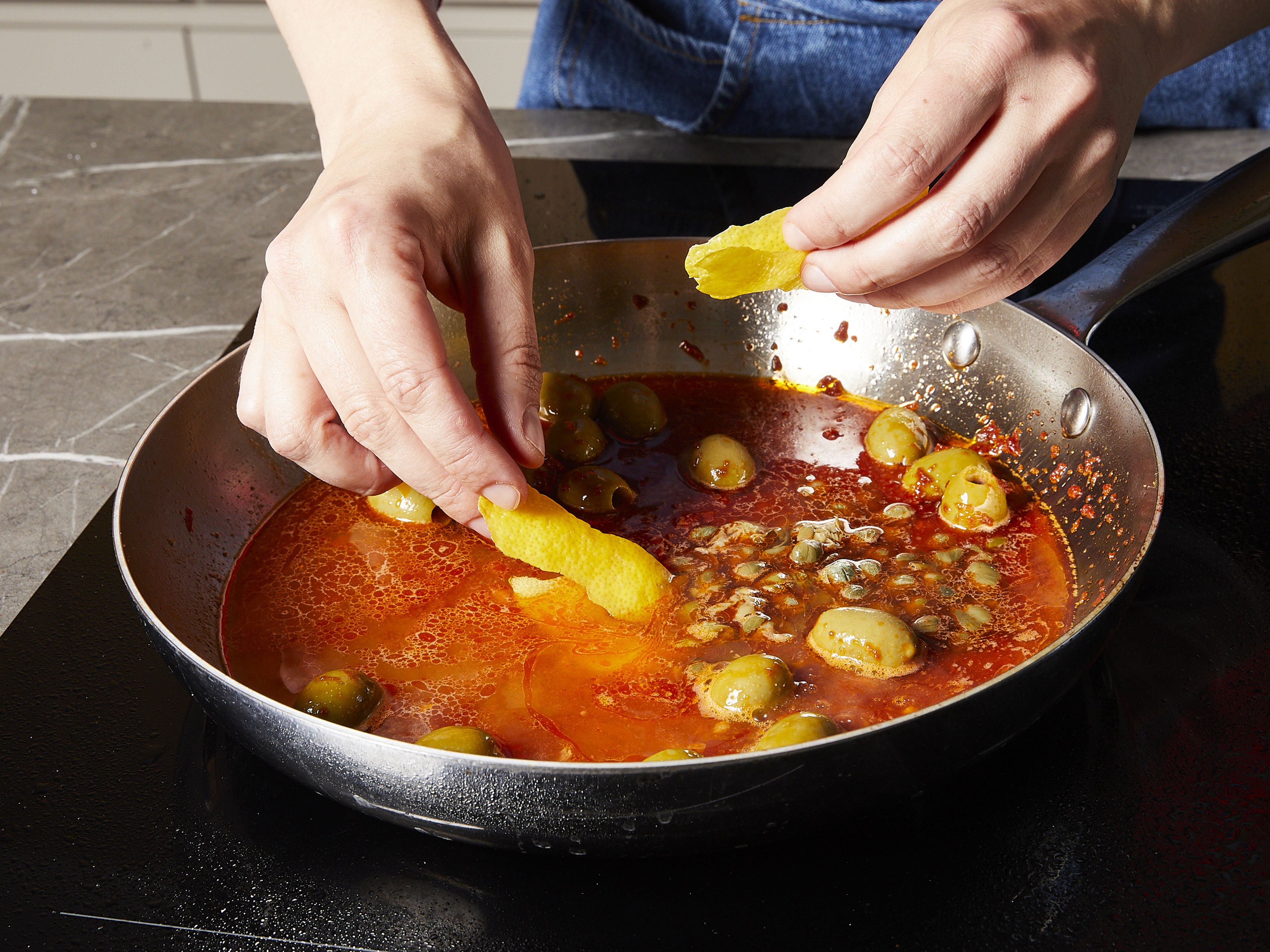 Add garlic and anchovies to the pan and sauté for approx. 2–3 min. Add chili flakes, tomato paste and sauté for another 2–3 min. Then, add olives, white wine, capers, bay leaves, lemon zest, and tomatoes to the pan. Bring to a simmer and leave it uncovered. Stir occasionally for approx. 3–5 min.