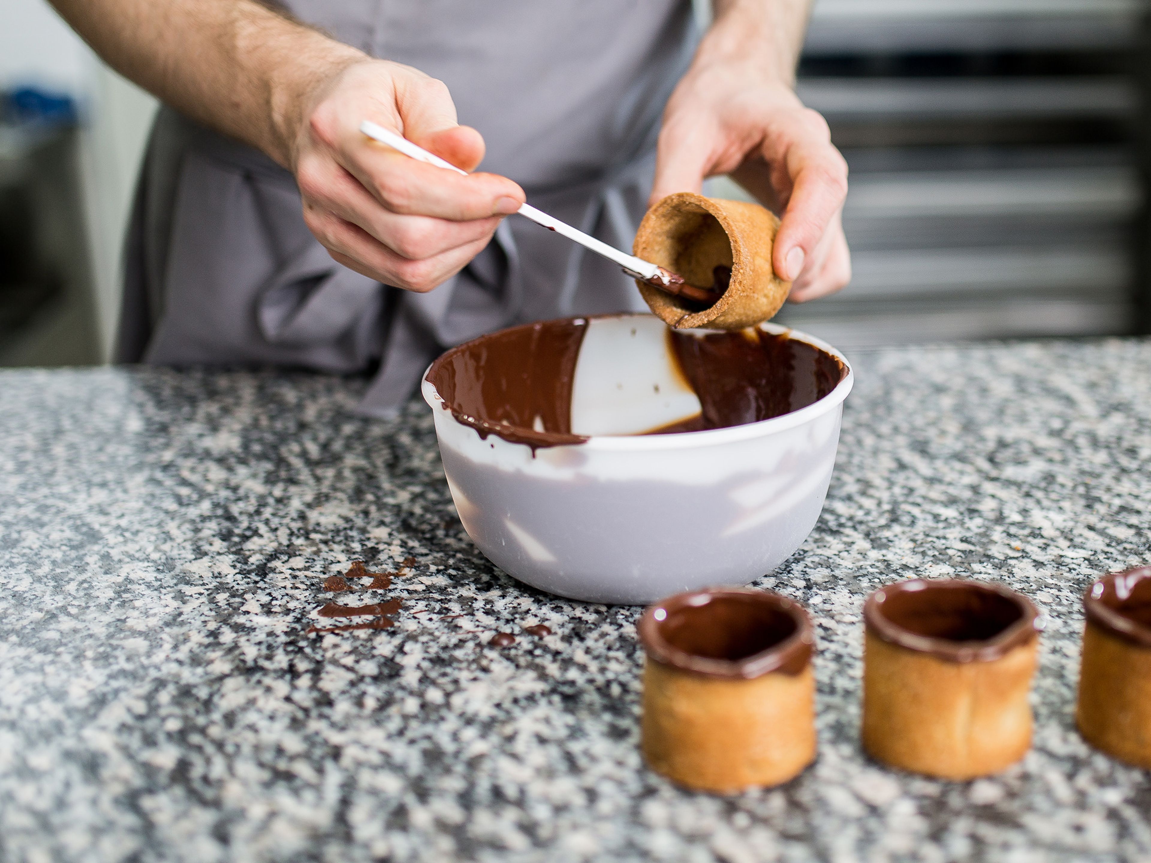 Sobald die Schokolade auf 32°C abgekühlt ist, das Innere der Cookie Cups zweimal damit glasieren. Für den Rand jeden Cup leicht in die Schokolade tauchen und die überschüssige Schokolade abtropfen lassen. Die Cups nach Belieben direkt mit Schokoladenstreuseln oder gehackten Mandeln verzieren. Sobald die Schokolade ausgehärtet ist kann der Rand auch mit essbarem Goldstaub verziert werden. Für ca. 20 Min. im Kühlschrank kaltstellen.