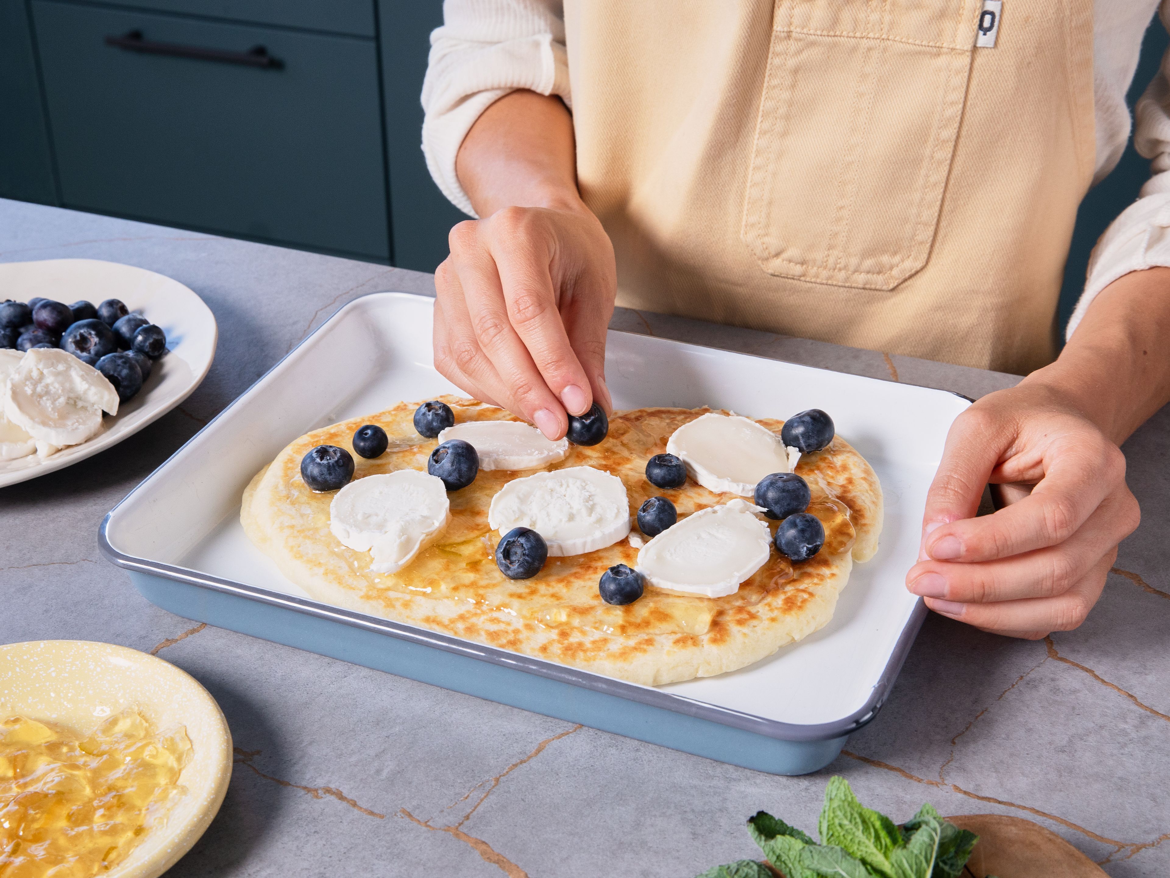 Blaubeeren waschen und einige zum Garnieren beiseitestellen. Ziegenkäse in Scheiben oder Stücke schneiden. Den Teig von der Pfanne auf ein mit Backpapier ausgelegtes Backblech setzen. Die Marmelade auf dem Fladenbrot verteilen. Mit Ziegenkäse und dem Großteil der Blaubeeren belegen, mit etwas Olivenöl besprenkeln und im vorgeheizten Backofen ca. 10 Min. backen. Mit frischer Minze, grobem Meersalz und dem Rest der Blaubeeren garnieren und servieren.