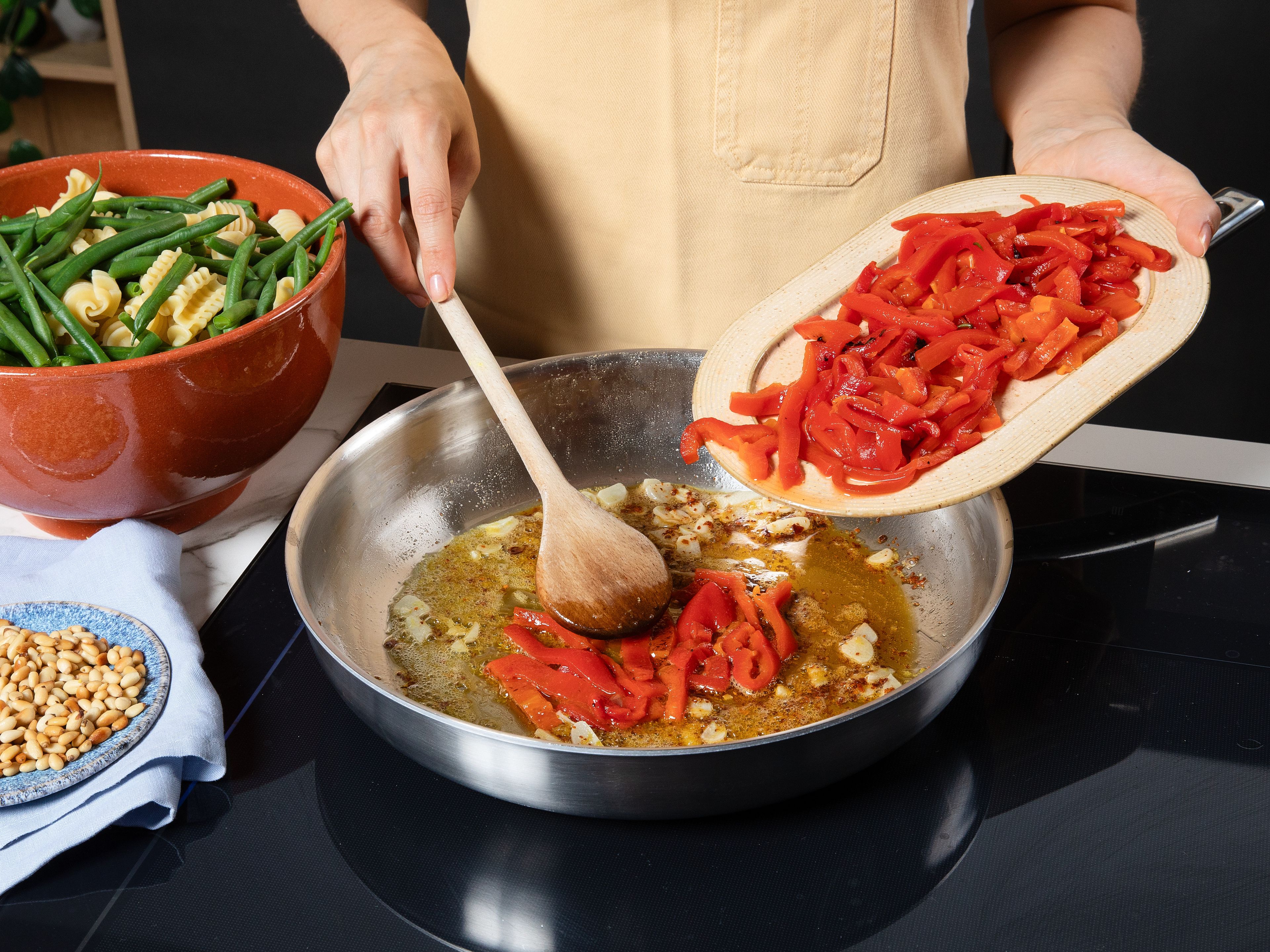 Die gerösteten Paprika abgießen und in Streifen schneiden. Den Knoblauch schälen und in feine Scheiben schneiden. Die Zitrone abreiben und auspressen. Die Pinienkerne in einer großen Pfanne goldbraun rösten und herausnehmen. In derselben Pfanne Olivenöl erhitzen, den Knoblauch zusammen mit den Chiliflocken und der Zitronenzeste hineingeben und kurz anschwitzen, bis es duftet. Die Paprikastreifen, Bohnen und Pasta hinzugeben und vorsichtig vermengen, bis alles gleichmäßig mit dem aromatischen Öl überzogen ist.