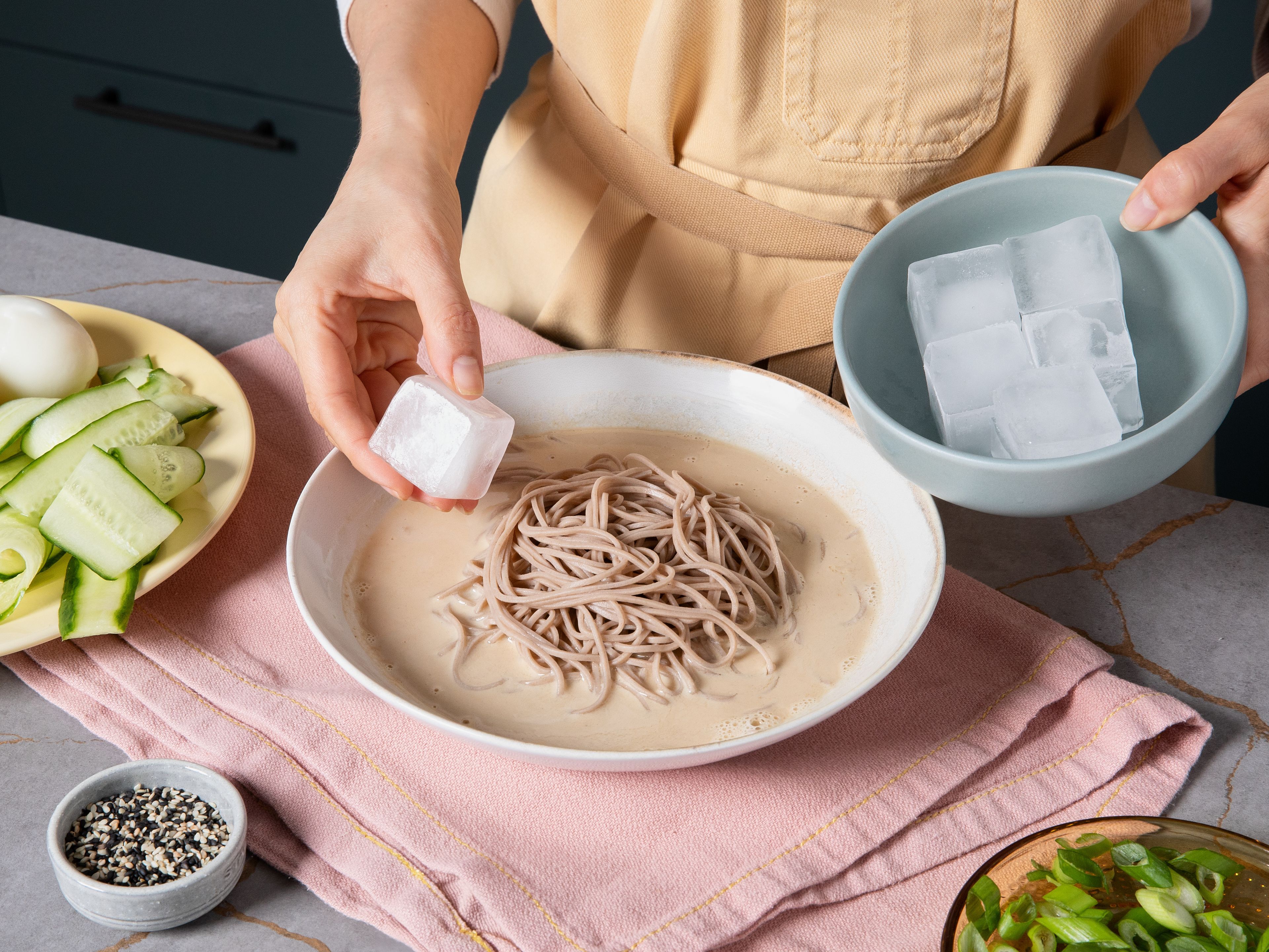 Gekühlte Nudeln in die Teller zur Tahini-Sojamilch-Brühe geben. Eiswürfel hinzufügen und mit Gurke, halbiertem Ei und nach Belieben weiteren Toppings belegen. Zum Schluss mit Chili Crisp beträufeln und Frühlingszwiebeln und Sesam bestreuen.