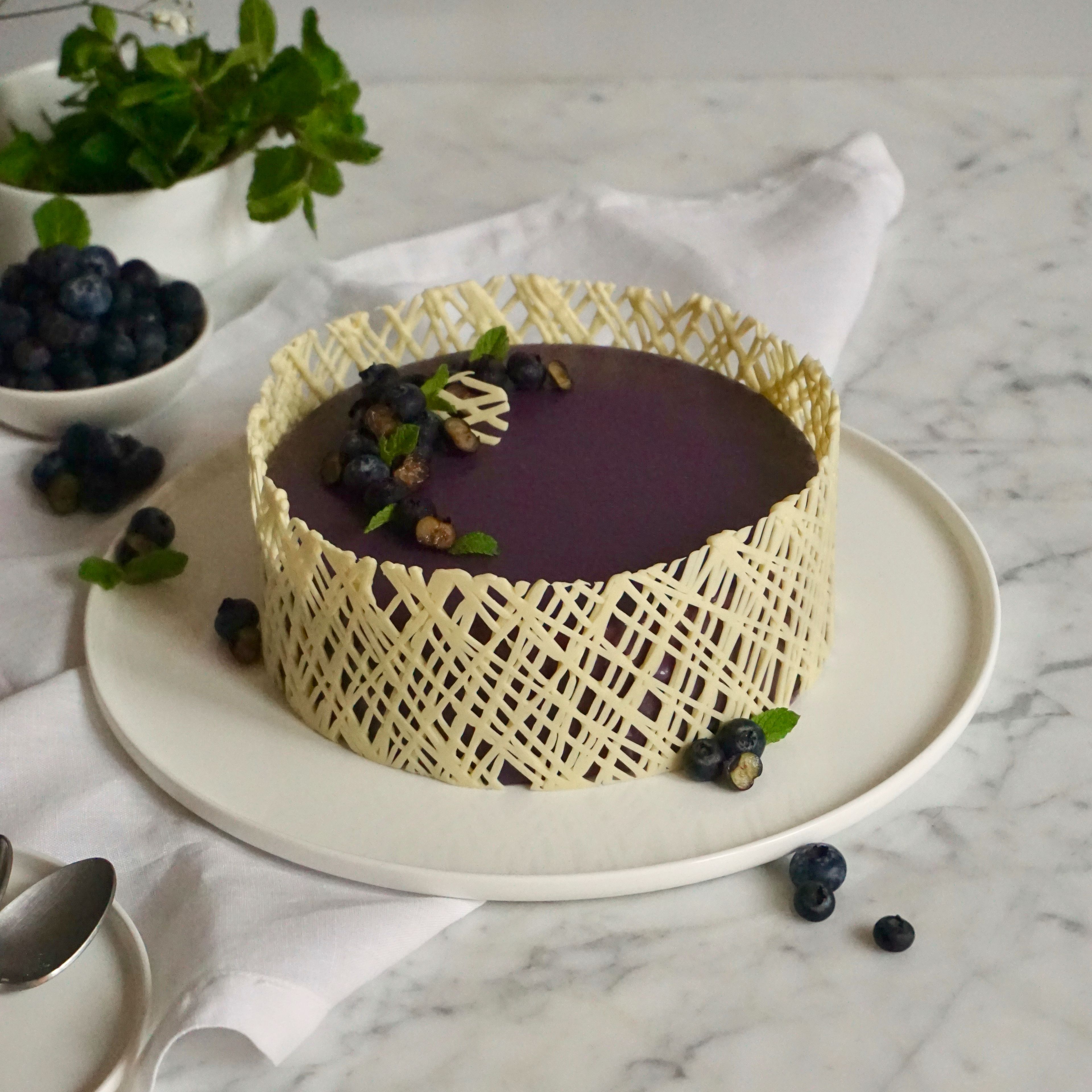 Torte aus dem Tiefkühler nehmen und aus dem Ring lösen. Torte auf eine kleine umgedrehte Box stellen. Glasur in Mikrowelle verflüssigen (ca. Körpertemperatur). Flüssige Glasur über die gefrorene Torte gießen. Den unteren Rand mit einer Winkelpalette abziehen und die Torte auf eine Tortenplatte setzen. Torte mit Blaubeeren und Minzblättern garnieren. Ring aus Schokolade von oben um das Entremet legen. Das Entremet nun für ca. 4 Stunden im Kühlschrank auftauen lassen.