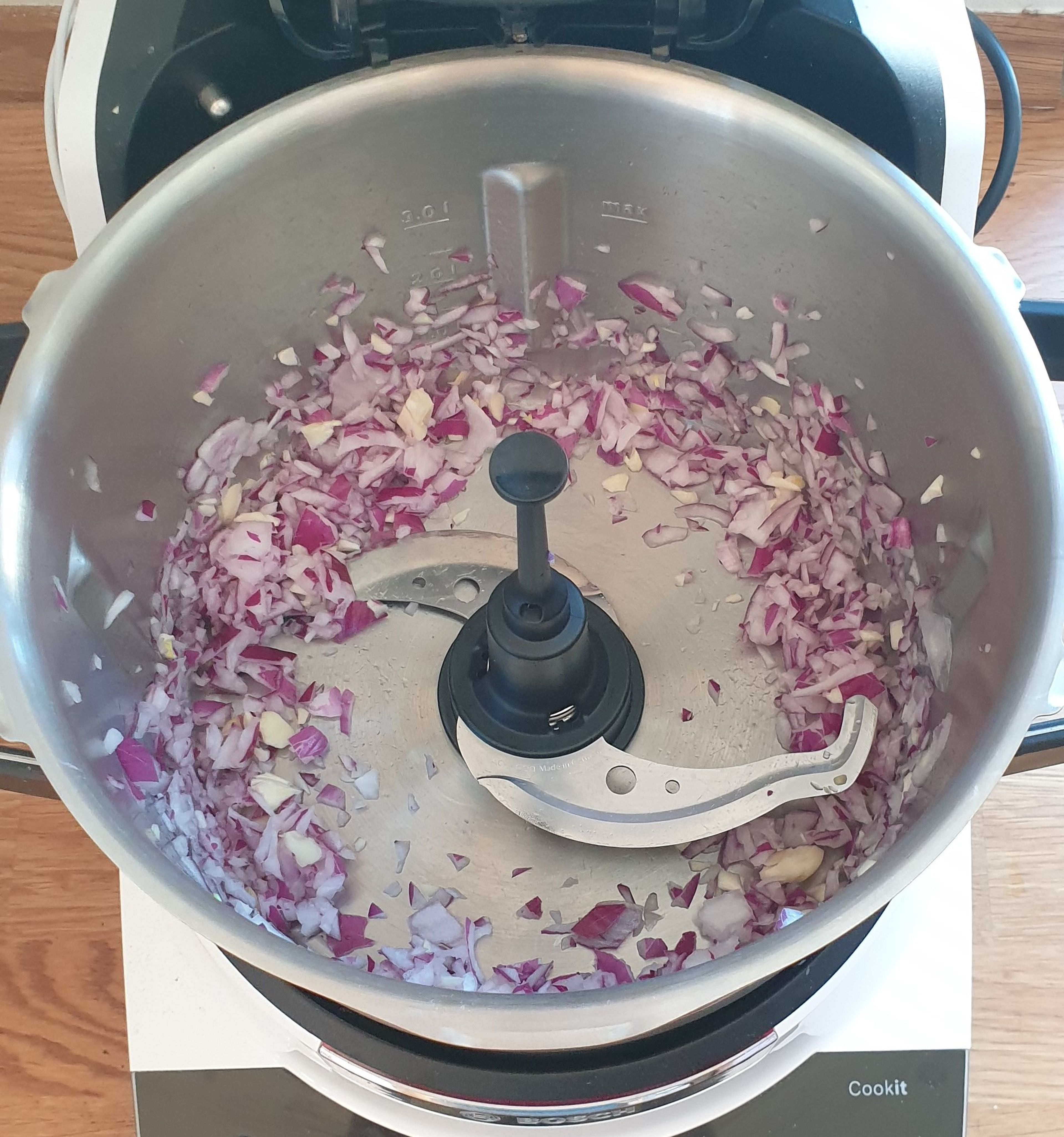 Das Universalmesser in den Cookit einsetzen. Die Zwiebel grob schneiden und mit den beiden Knoblauchzehen bei geschlossenem Deckel und eingesetztem Messbecher zerkleinern (Universalmesser | Stufe 14 | 5 Sekunden).