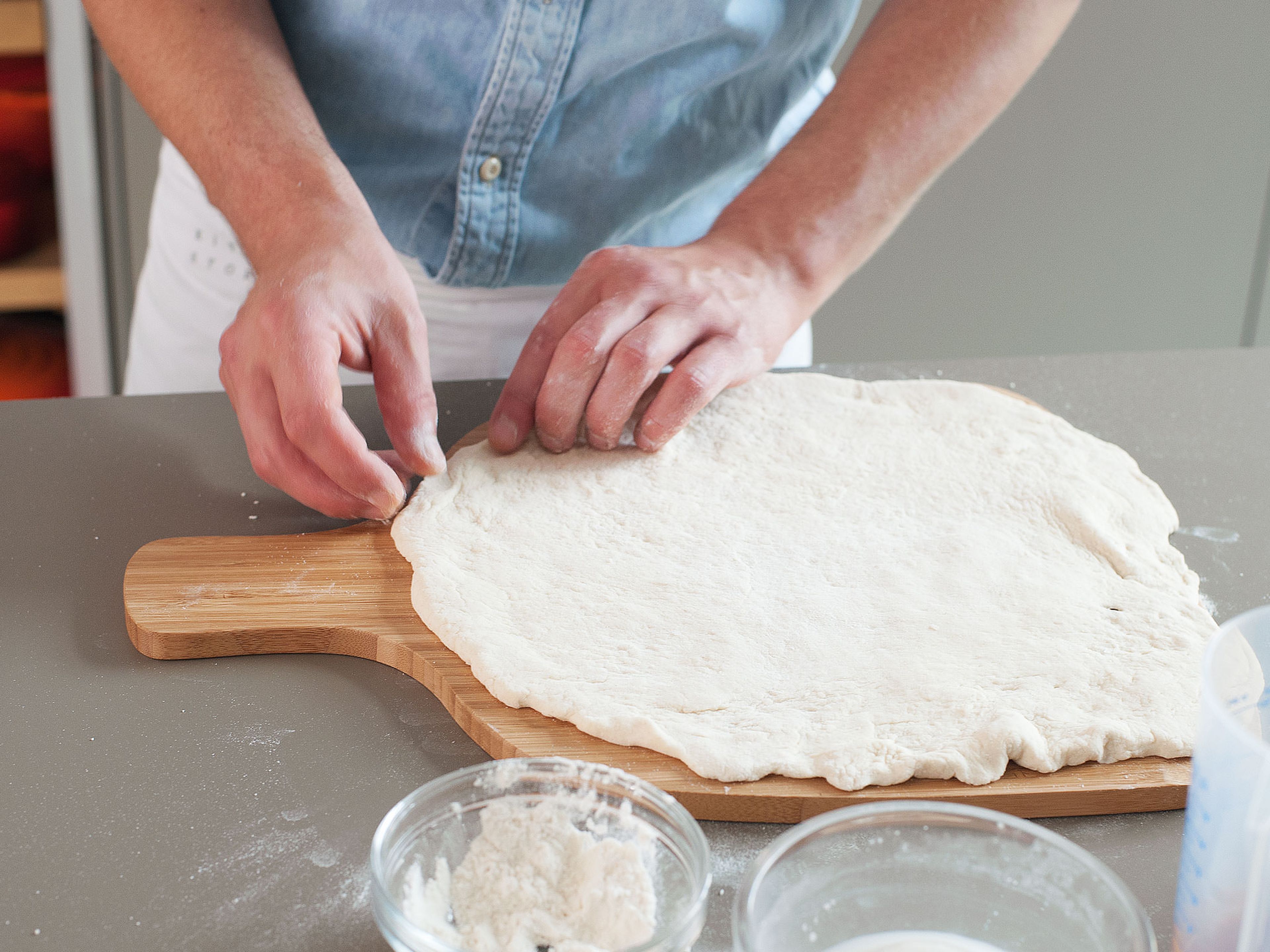 Teig aus der Schüssel nehmen und auf einer mit Mehl bestäubten Arbeitsfläche ungefähr auf die Größe des Pizzasteins ausbreiten. Anschließend auf den Pizzaheber legen.