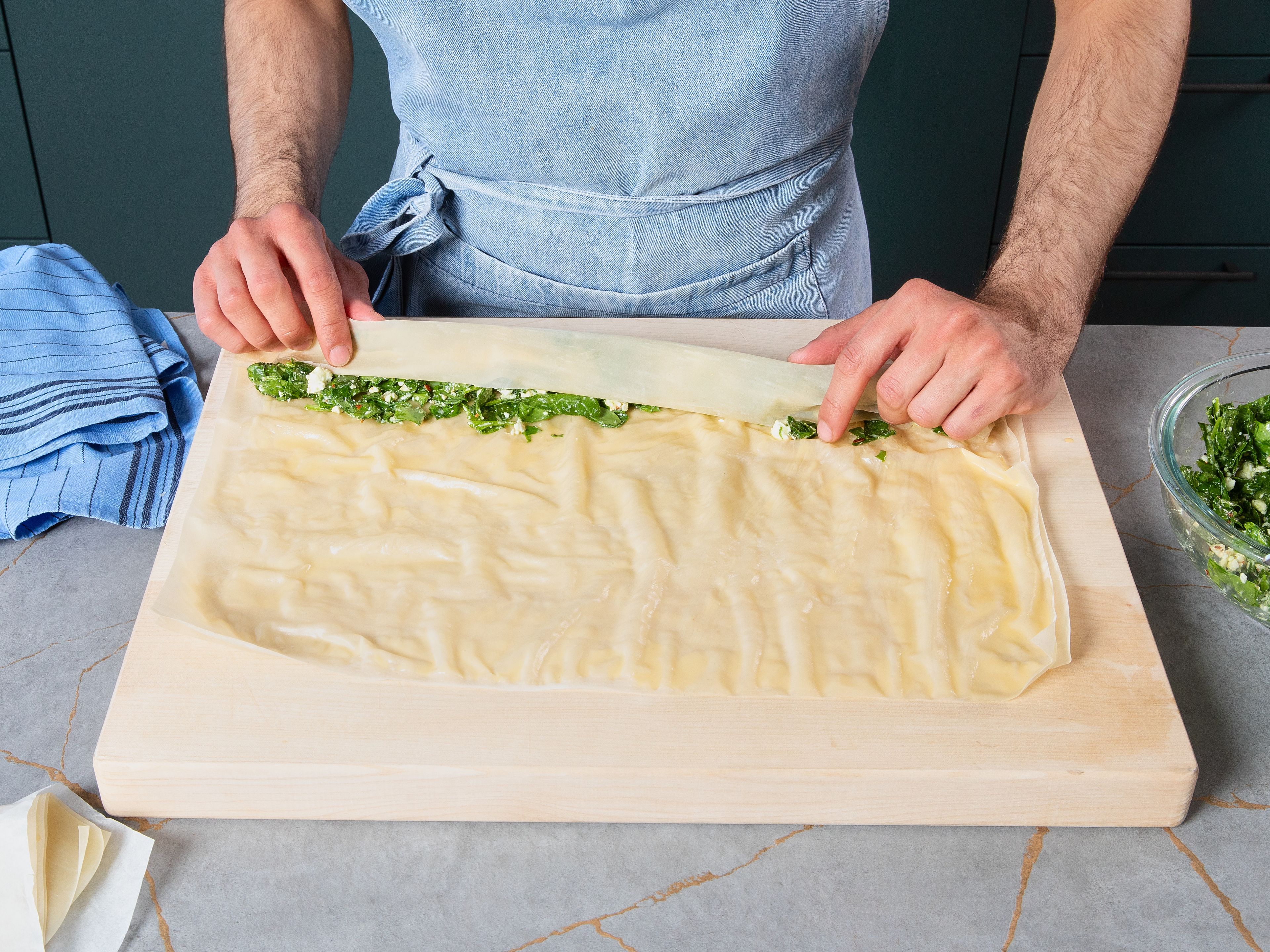 For the filling, roughly chop spinach and scallion. Crumble feta. Then, combine spinach, scallion, feta, chili flakes, oil, salt, and pepper in a bowl. Grease a springform pan and line it with parchment paper. Place filling along the long side of the dough. Roll the filling in from the same long side.