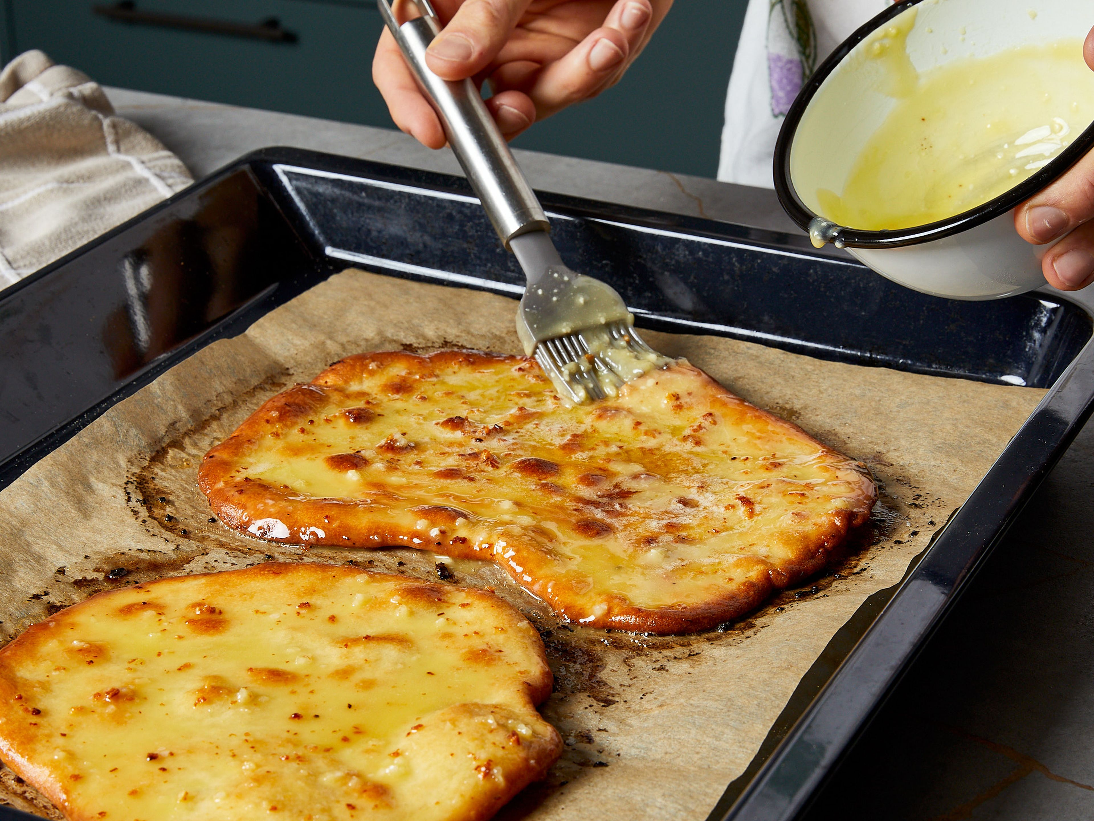 Combine oil and butter in a bowl until smooth. Crush the garlic and add most of it to the oil-butter mixture. Season with salt and pepper. Generously brush half of the garlic mixture over the Lángos pieces and bake for approx. 20 min., until golden brown. After baking, brush them with the remaining garlic mixture. For the toppings, mix sour cream with the rest of the crushed garlic and spread it over each piece. Finish with shredded mozzarella, crispy bacon bits, sliced onion, and chives to your liking.