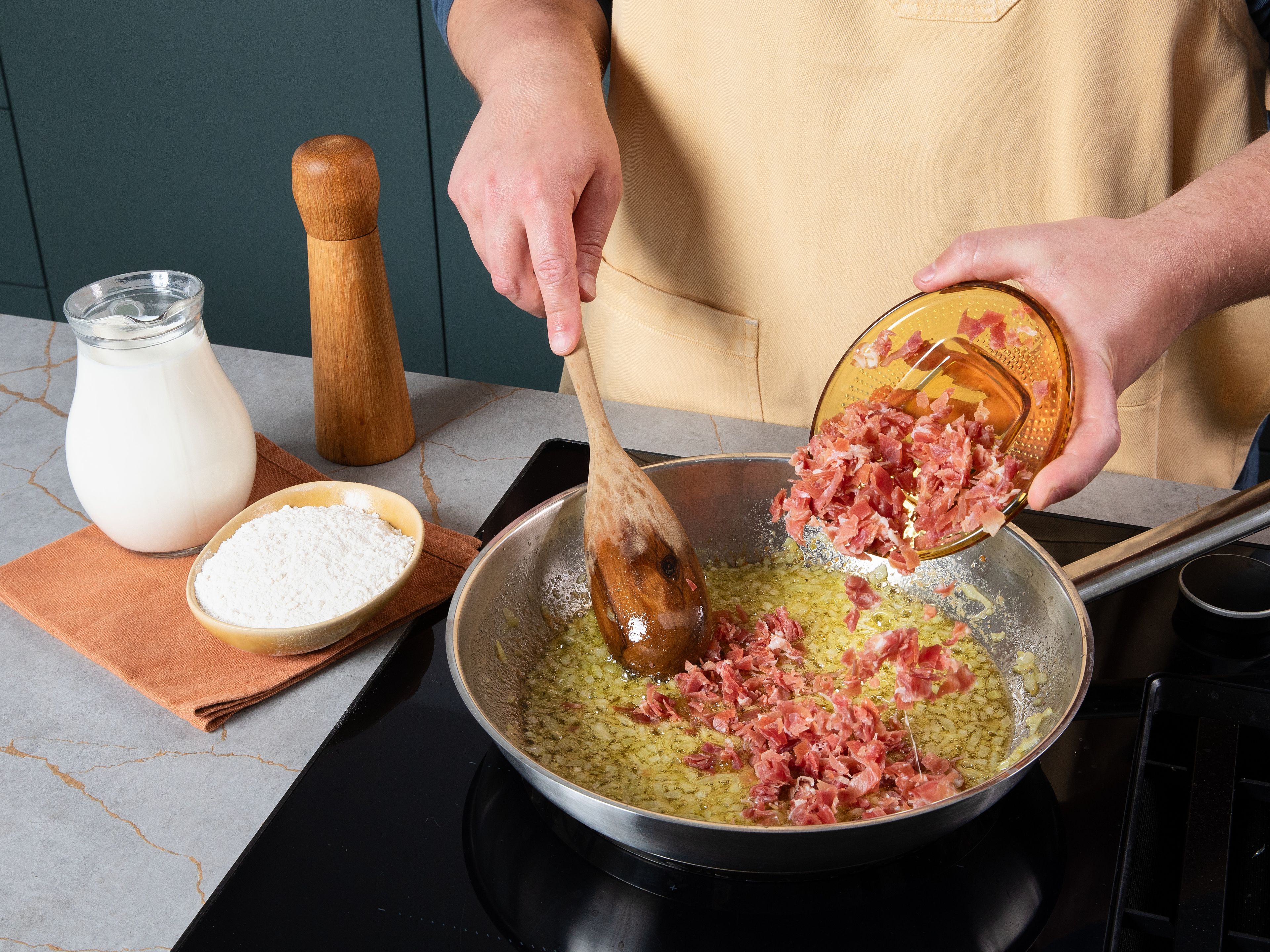 Zwiebel fein würfeln und Serrano-Schinken fein hacken. Butter und Olivenöl in einem Topf erhitzen und die Zwiebel darin glasig anschwitzen. Serrano-Schinken hinzufügen und kurz mitbraten, dann Mehl einrühren und kurz mitrösten. Anschließend nach und nach unter ständigem Rühren die Milch hinzugeben, Muskatnuss einrühren und köcheln lassen, bis eine dicke, cremige Masse entsteht. Die Masse in eine eingefettete Form füllen, abkühlen lassen, mit Frischhaltefolie abdecken und für mindestens ca. 4 Std. (am besten über Nacht) kalt stellen.