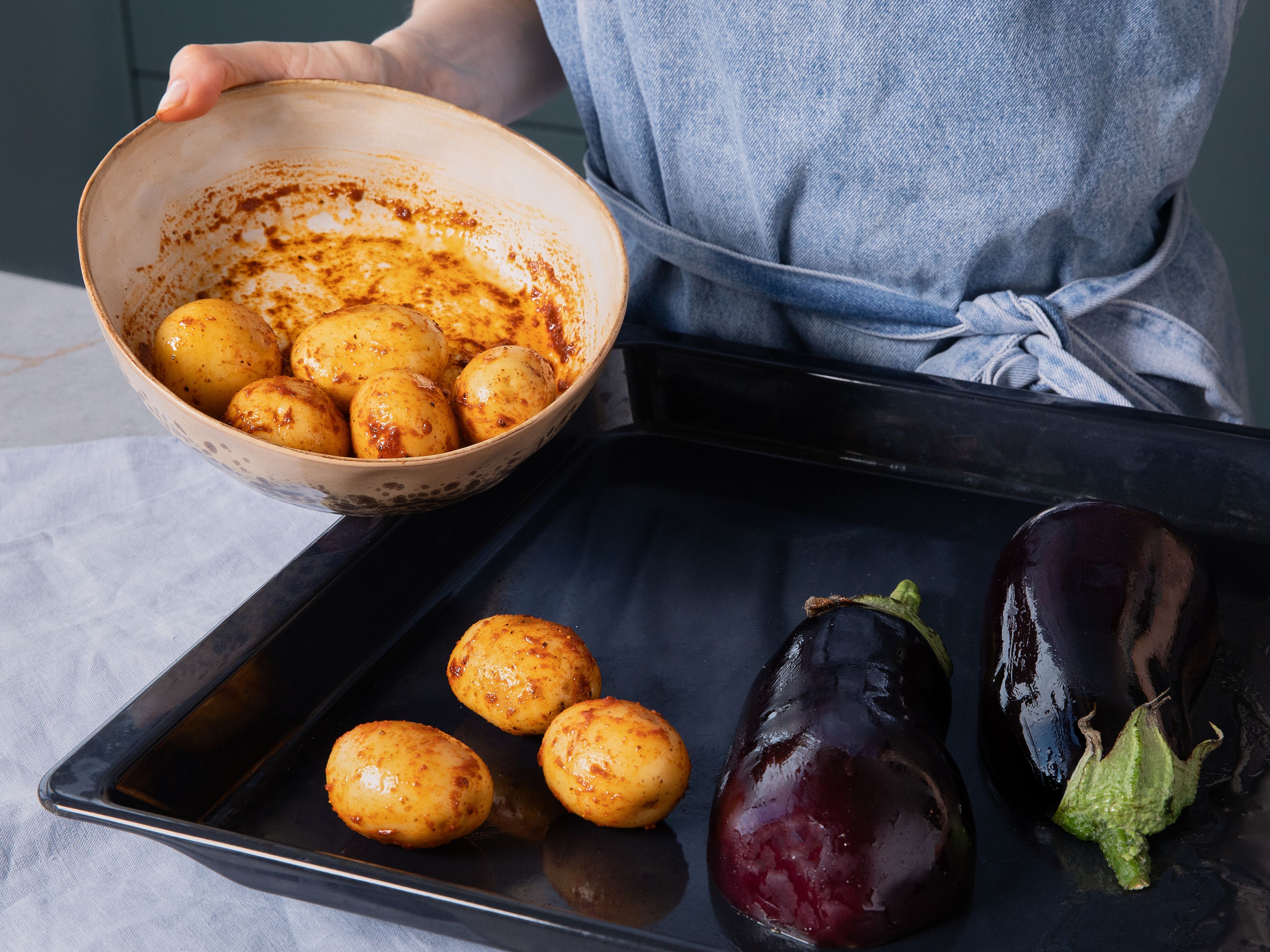 Heize den Backofen auf 200 °C (Ober-/Unterhitze) vor. Die Aubergine halbieren, großzügig mit Olivenöl bestreichen, salzen und mit der Schnittfläche nach unten auf ein Backblech legen. Drillinge je nach Größe halbieren und in einer Schüssel mit Olivenöl, Harissa, Salz und Pfeffer vermengen. Kartoffeln zu den Auberginen auf das Backblech geben. Alles ca. 25 Min., rösten, bis die Auberginen innen weich und unten goldbraun sind und die Kartoffeln goldbraun und knusprig sind.