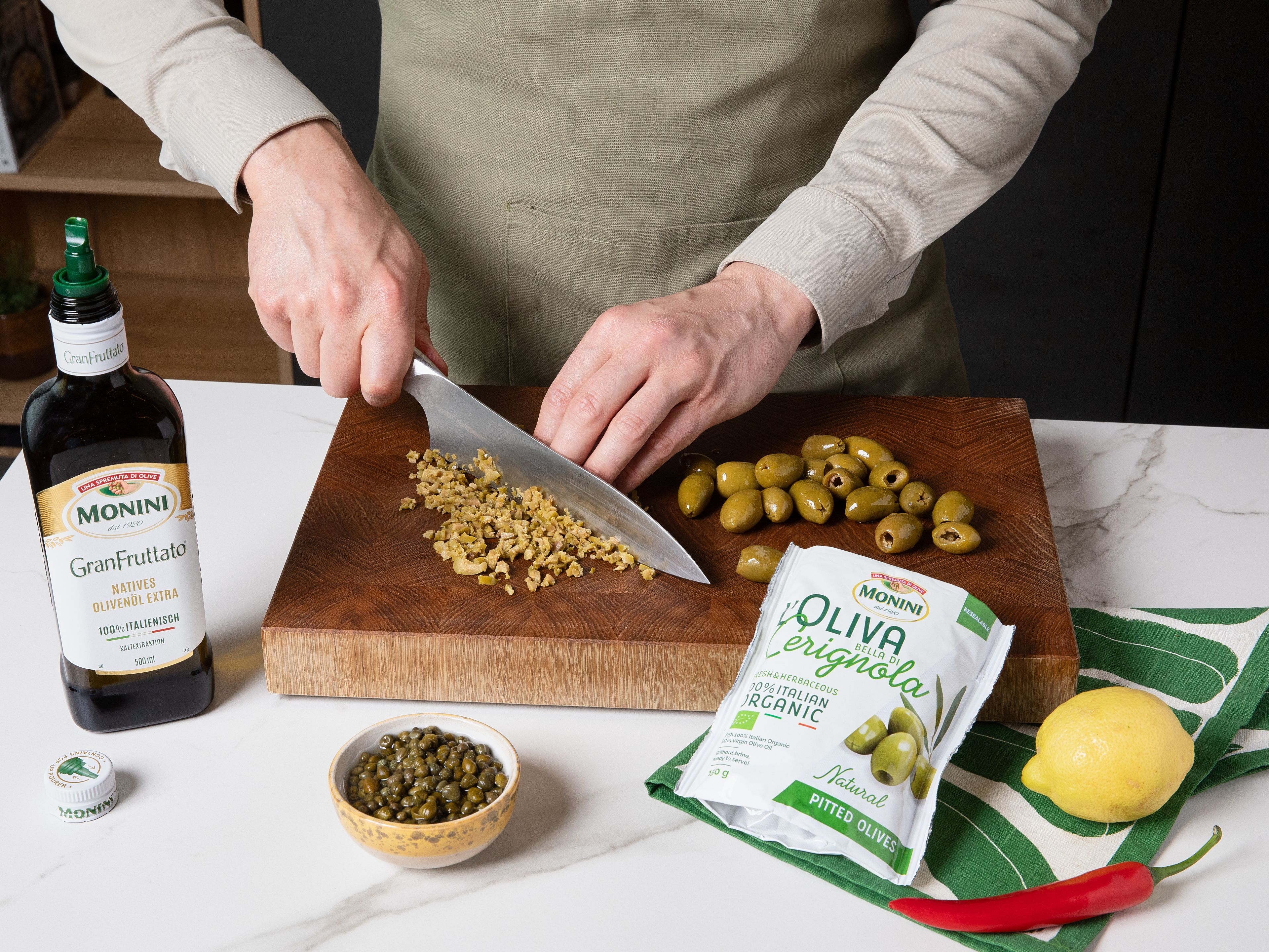 Oliven fein hacken. Die Zitrone abreiben und auspressen. Kapern, Petersilie und Chili fein hacken. Alles mit 3 EL Monini Gran Fruttato Olivenöl vermengen und mit Salz und Pfeffer abschmecken.