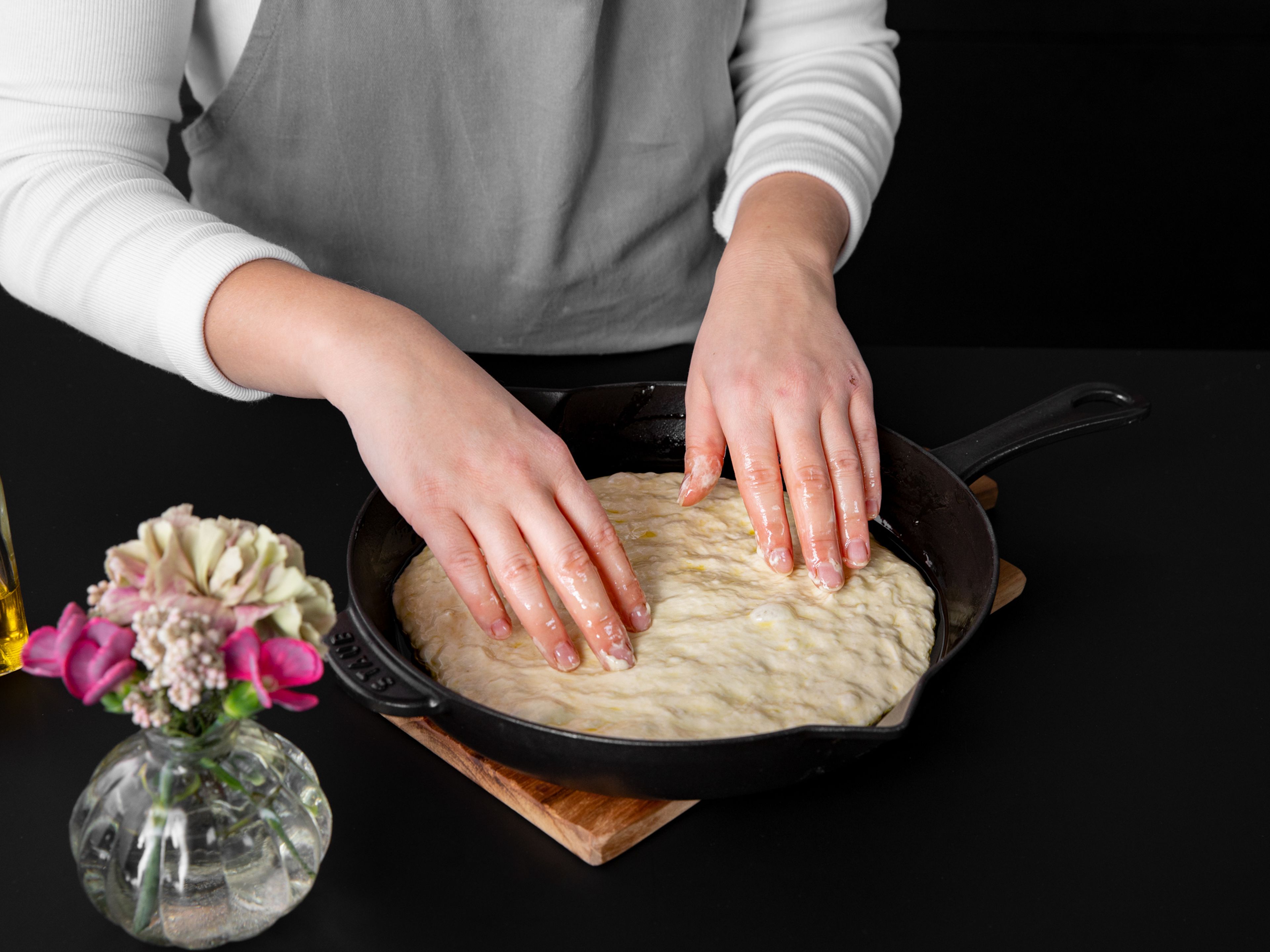 Den Ofen auf 250°C vorheizen. Gib etwas Olivenöl in die Gusspfanne. Fette auch die Seiten der Pfanne mit einem Gummispatel oder deinen Händen ein. Gib eine Hälfte des Teiges in die Pfanne. Falte den Teig einige Male von den Seiten zur Mitte hin ein und drehe in dann um, sodass die gefaltete Seite an der Unterseite ist. Verteile den Teig gleichmäßig in der Pfanne, indem du ihn mit deinen Fingern in Form drückst. Für ca. 1 Stunde ruhen lassen.