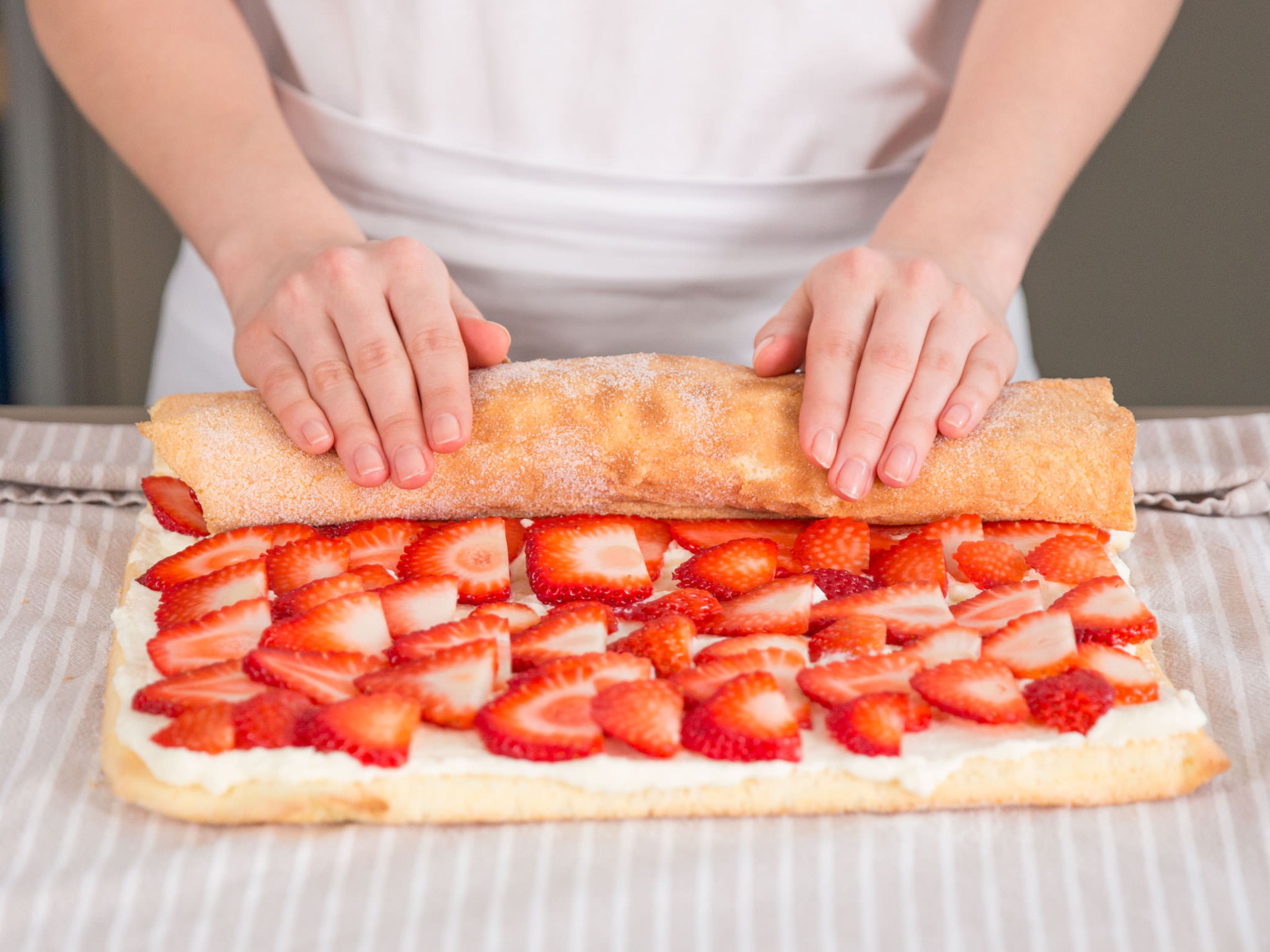 Sicherstellen, dass der Kuchen komplett abgekühlt ist. Ca. zweidrittel der Sahnefüllung auf dem Kuchen verteilen, gefolgt von einer gleichmäßigen Schicht Erdbeeren. Anschließend mit Hilfe des Handtuchs nach vorne zusammenrollen.