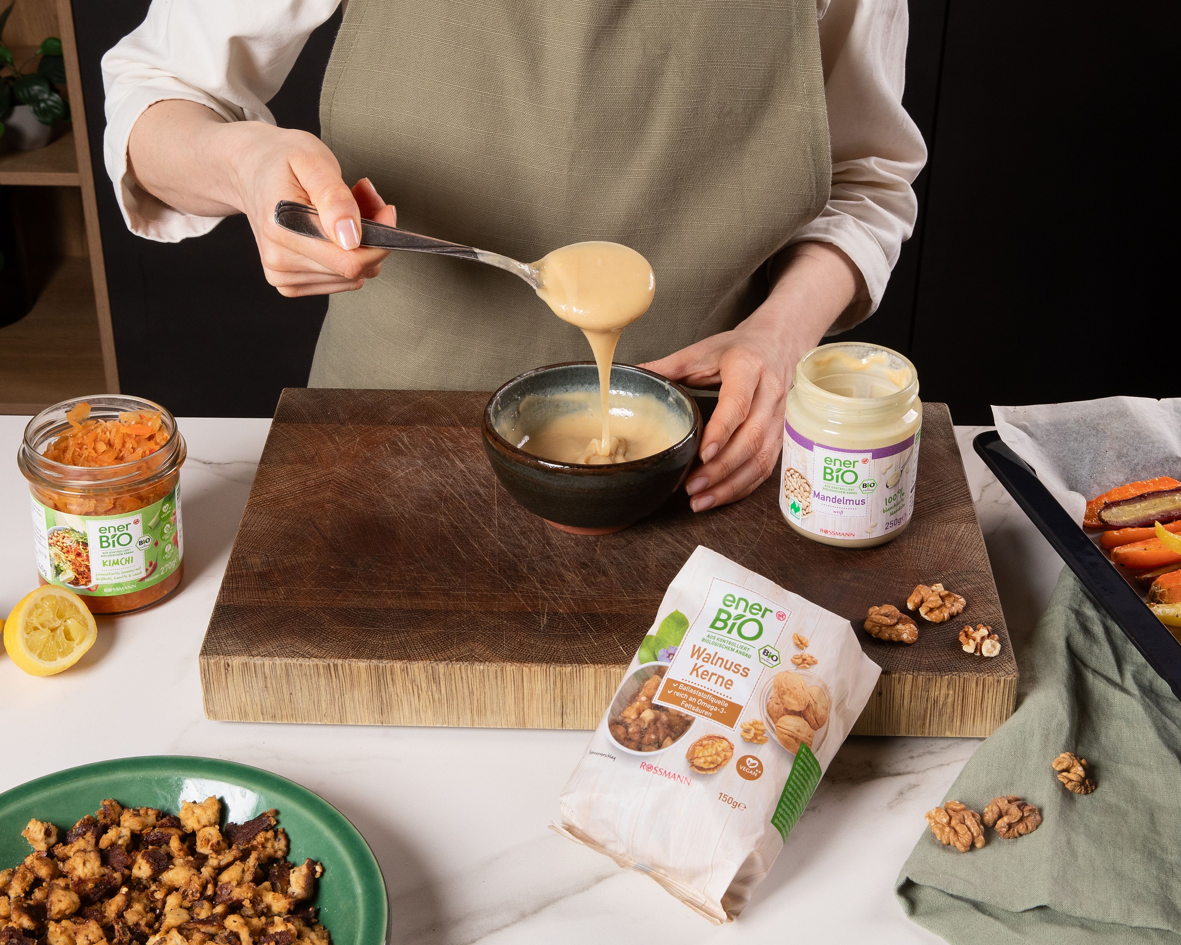 Das enerBiO Kimchi abtropfen. Die enerBiO Walnusskerne hacken. In einer kleinen Schüssel das enerBiO weiße Mandelmus mit Zitronensaft verrühren, bis ein cremiges Dressing entsteht. Mit Salz, enerBiO Pfeffer und enerBiO Agavendicksaft abschmecken und bei Bedarf mit etwas Wasser verdünnen. Den Koriander grob hacken. Das geröstete Gemüse aus dem Ofen nehmen und mit dem geschnittenen Kimchi vermengen. Auf Tellern anrichten, mit dem Mandelmus-Dressing beträufeln und mit dem Tofu-Crumble, gehackten Walnüssen und frischem Koriander bestreuen. enerBiO Tofu Mandel-Nuss (AT-BIO-301); enerBiO Mandelmus weiß (DE-ÖKO-001); enerBiO Leinsamen geschrotet (DE-ÖKO-001); enerBiO Walnusskerne (DE-ÖKO-006); enerBiO Kimchi (DE-ÖKO-007), enerBiO Sesamöl geröstet (DE-ÖKO-006), enerBiO Agavendicksaft (DE-ÖKO-001), enerBiO Pfeffer (DE-ÖKO-006)