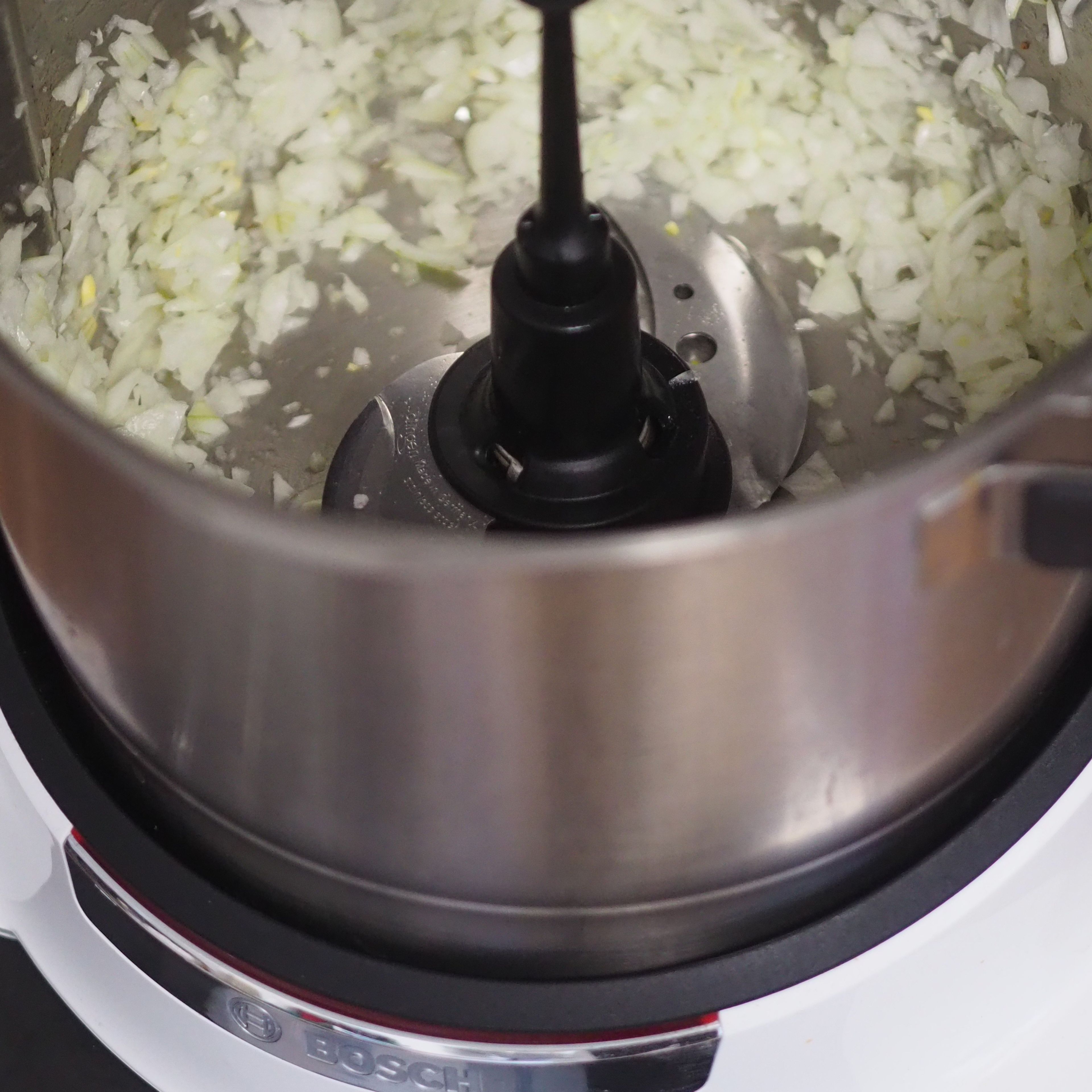 Die Zwiebeln schälen und halbieren. Das Universalmesser einsetzen. Beides in den Cookit geben, den Deckel schließen, den Messbecher einsetzen und zerkleinern (Universalmesser | Stufe 18 | 10 Sek.).
