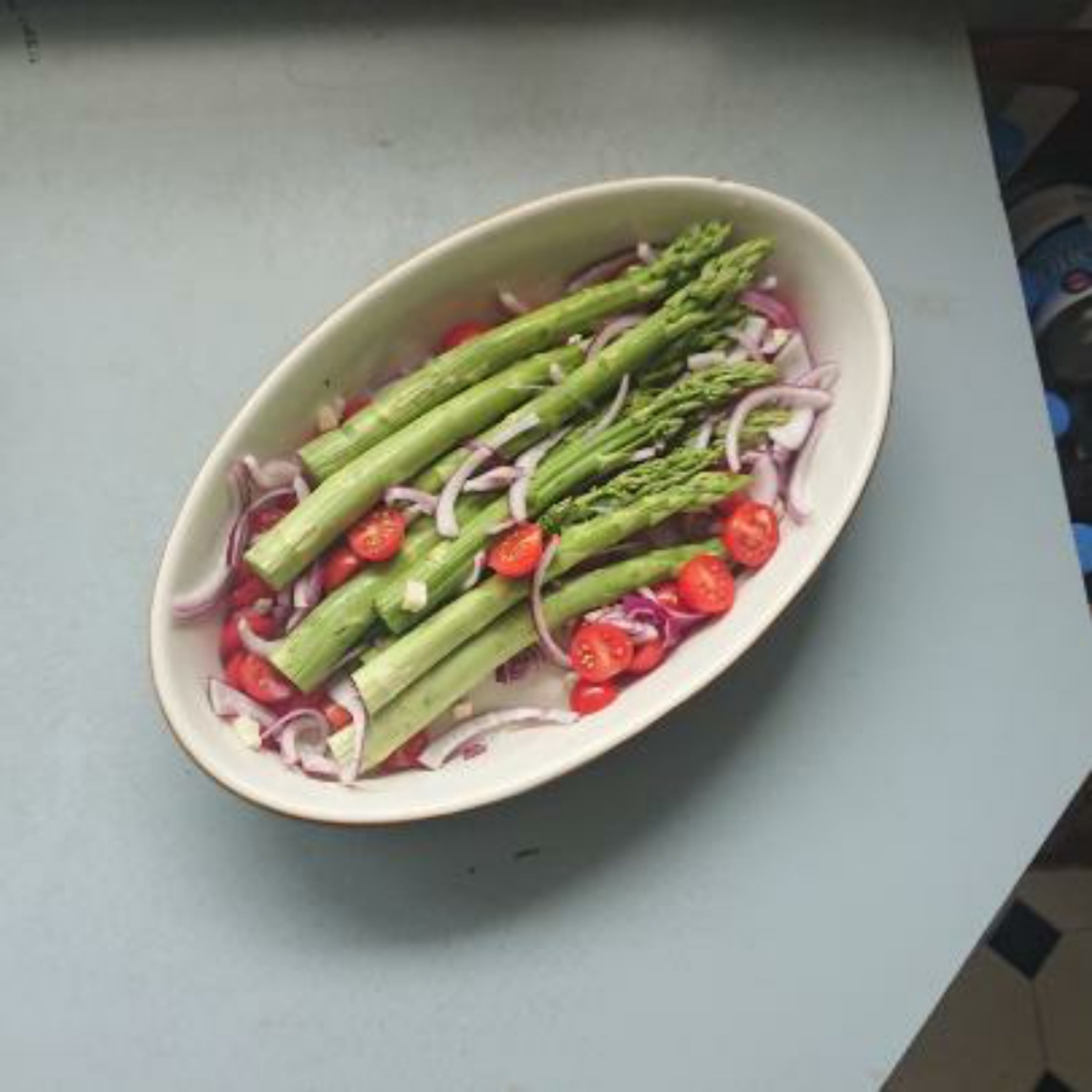 Die Cocktailtomaten waschen und halbieren. Zwiebel in Ringe schneiden und die Knoblauchzehen grob würfeln. Dann alles zum Spargel in die Form geben und vermischen