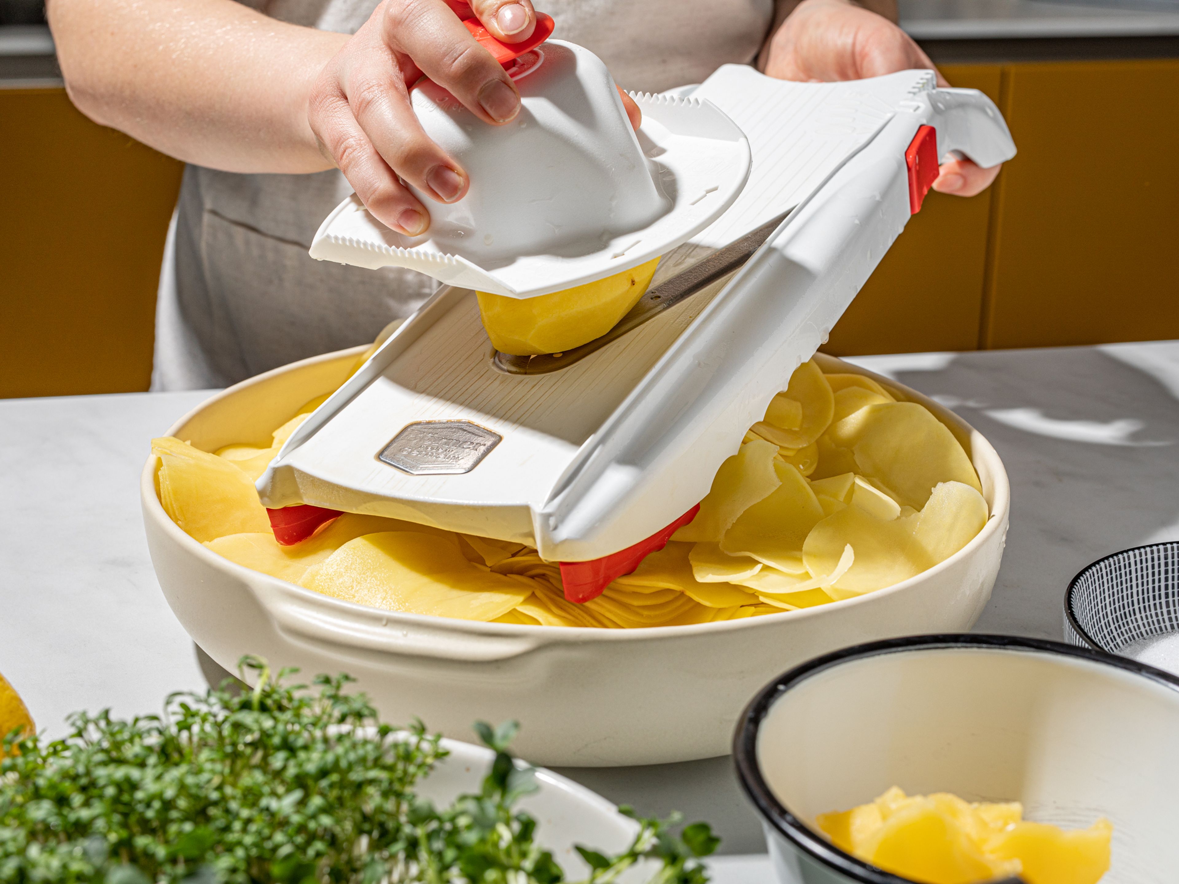 Den Butterschmalz in einem kleinen Topf erhitzen, bis er flüssig ist. Nun die Kartoffeln der Länge nach auf der dünnsten Stufe hobeln und in eine große Schüssel geben. Den Butterschmalz dazugeben und alles gut vermischen. Den Backofen auf 160°C vorheizen.