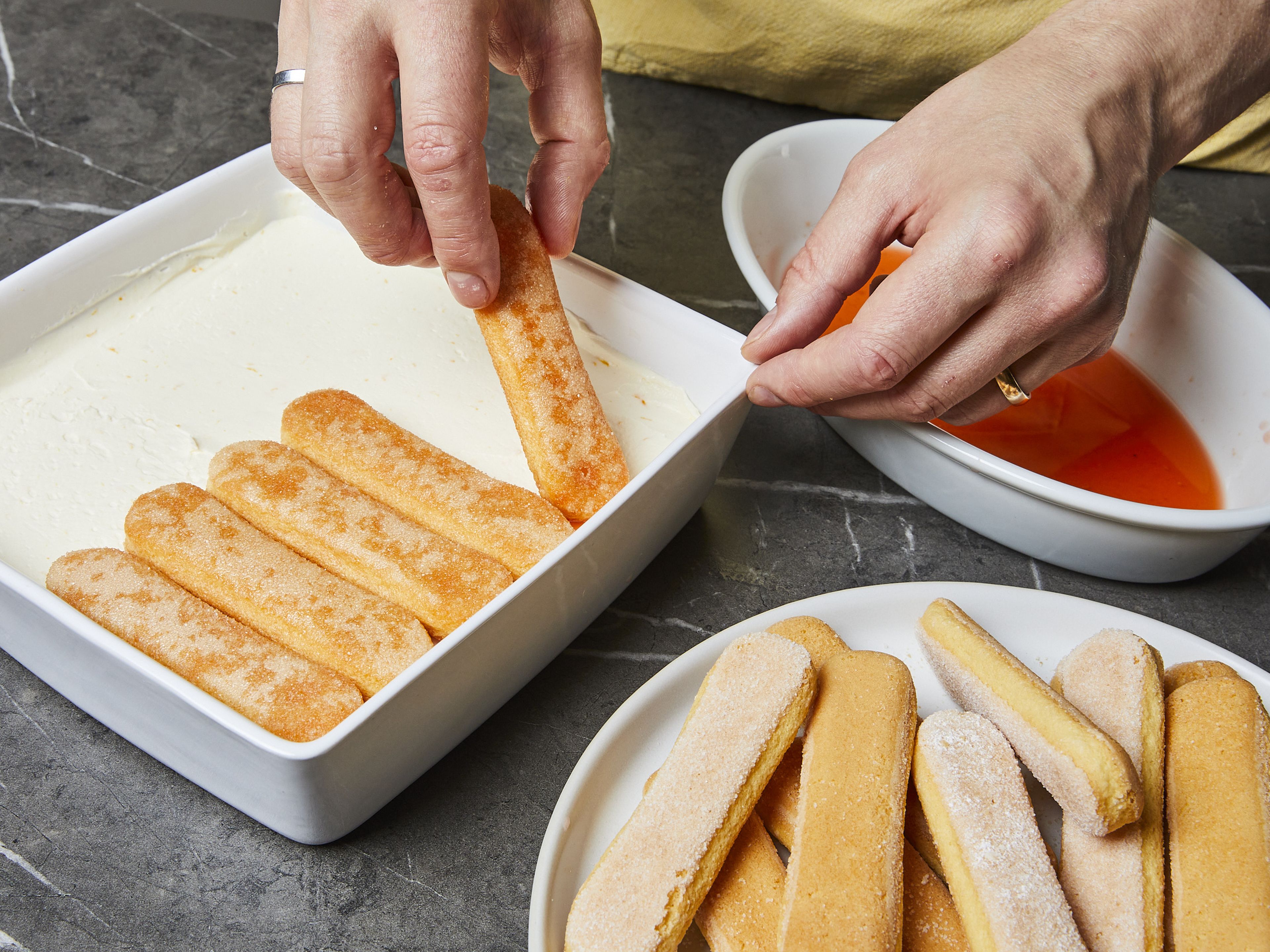 Briefly dip the ladyfingers one by one into the cooled syrup and cover the base of a baking dish. Spread a little less than half of the mascarpone cream on top and smooth out. Spread another layer of soaked ladyfingers on top and press down lightly. Spread the rest of the cream on top and smooth down as well. Stir the orange cream until smooth and pour over the top of the cream. Smooth out the orange cream and chill the tiramisu for at least 2–3 hrs. Garnish with orange slices and mint.