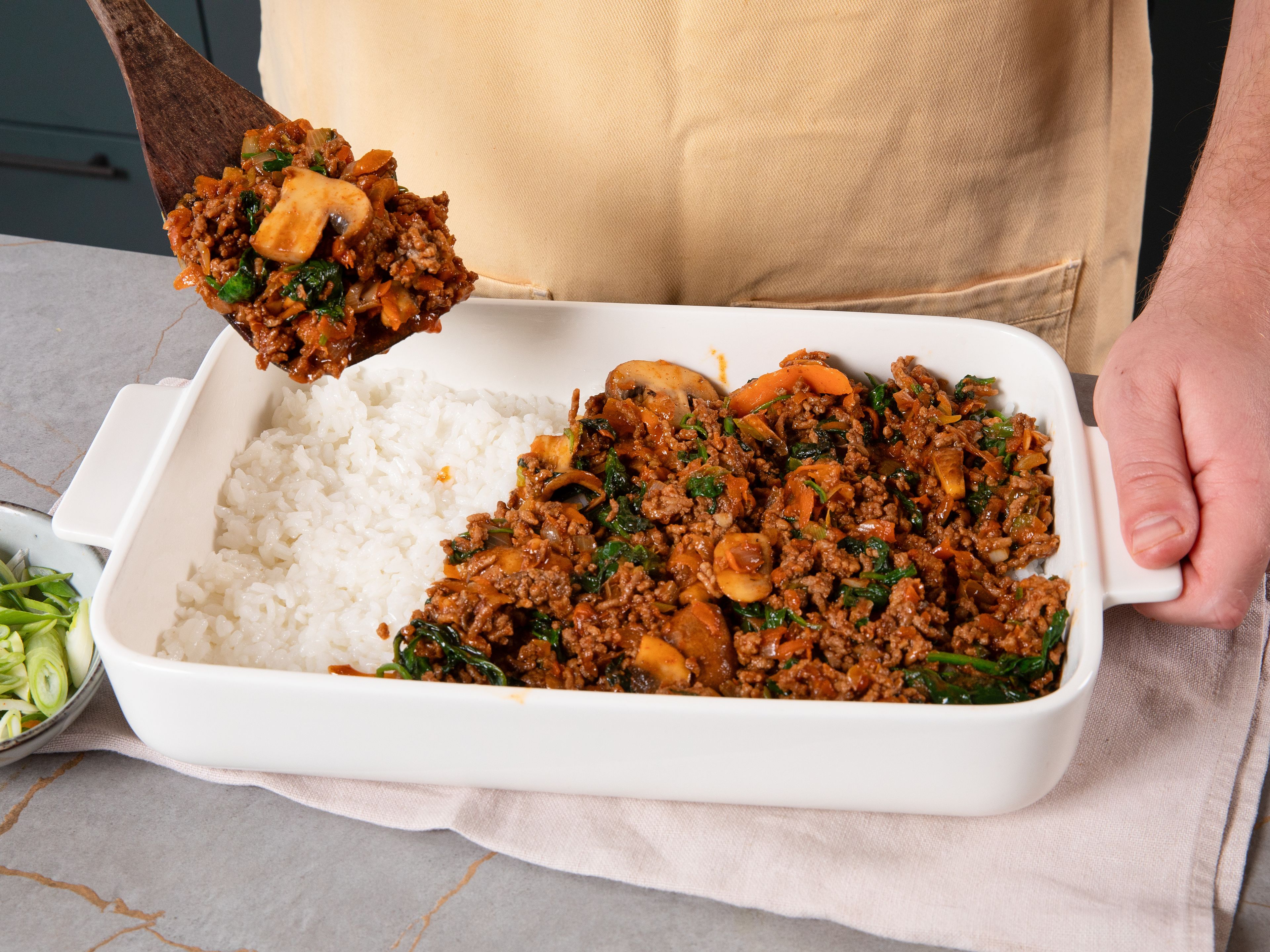 Das restliche Öl in derselben Pfanne erhitzen. Zwiebel, Knoblauch und Pilze hineingeben und ca. 2 Min. braten, bis die Pilze gebräunt sind. Spinat und Karotten hinzufügen und weitere ca. 2 Min. braten. Hackfleisch und Gochujang-Soße unterrühren und alles gut vermengen. Mit Salz und Pfeffer würzen.
In der gefetteten Auflaufform zuerst eine Schicht Klebreis verteilen. Anschließend die Hackfleischmischung darauf geben und gleichmäßig verstreichen.