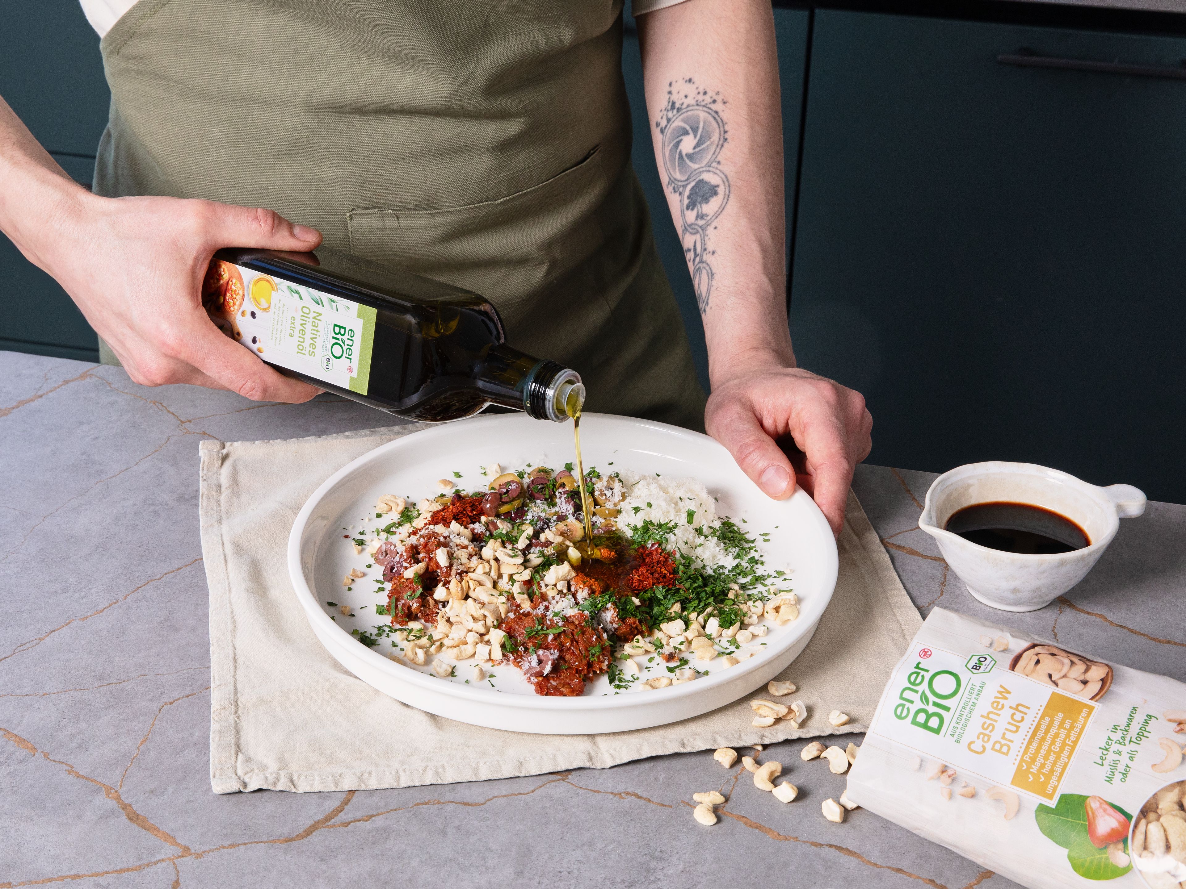 Das enerBiO Native Olivenöl extra und Balsamicoessig auf einen großen, tiefen Teller geben. Paprikapulver und Chiliflocken, die vorbereiteten Tomaten, Oliven, den Knoblauch, enerBiO Cashewbruch, die Petersilie und den geriebenen Parmesan dazugeben.