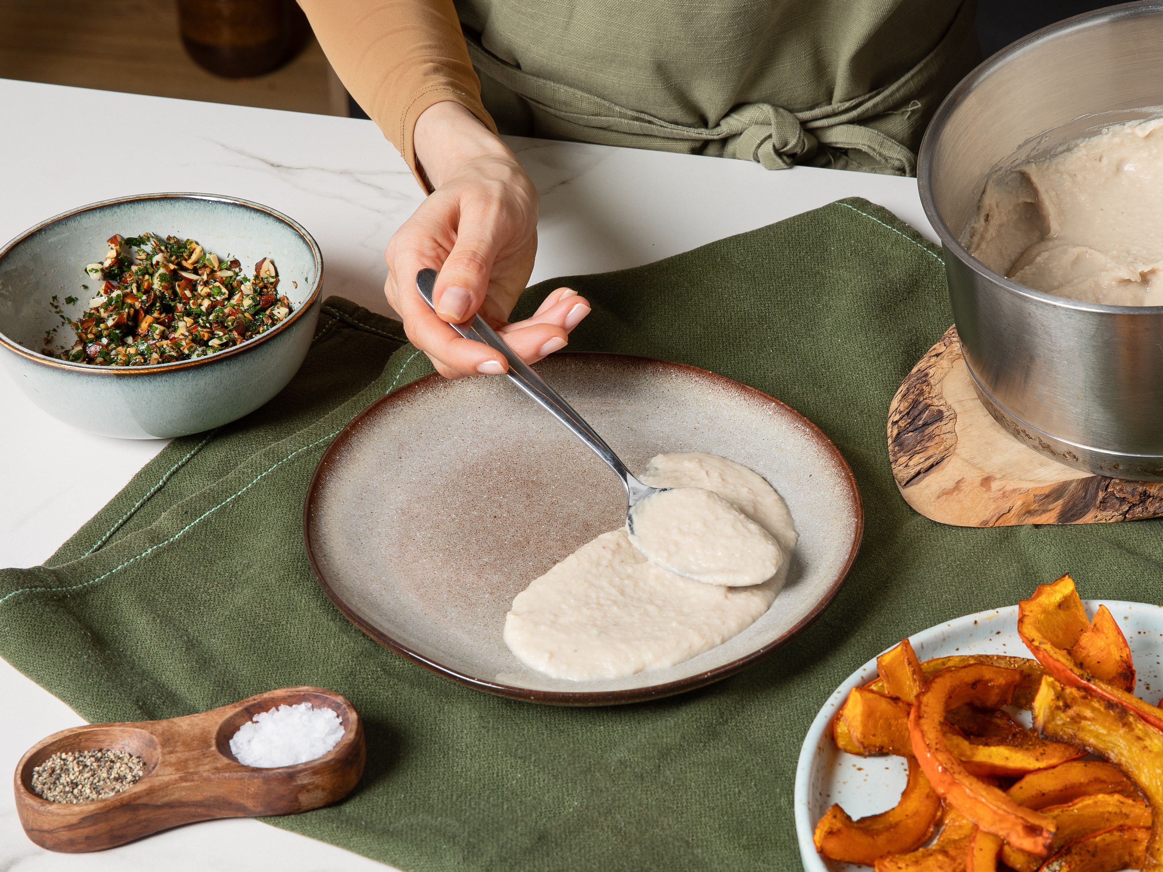Für die Gremolata die gehackte Petersilie zusammen mit Zitronenzeste, dem restlichen Knoblauch, Salz und etwas Olivenöl zu den gerösteten Mandeln in die Schüssel geben und vermengen. Das Bohnen-Mandel-Püree auf Tellern verstreichen und die gerösteten Kürbisspalten aus dem Ofen darauflegen. Mit der Gremolata garnieren und sofort servieren.