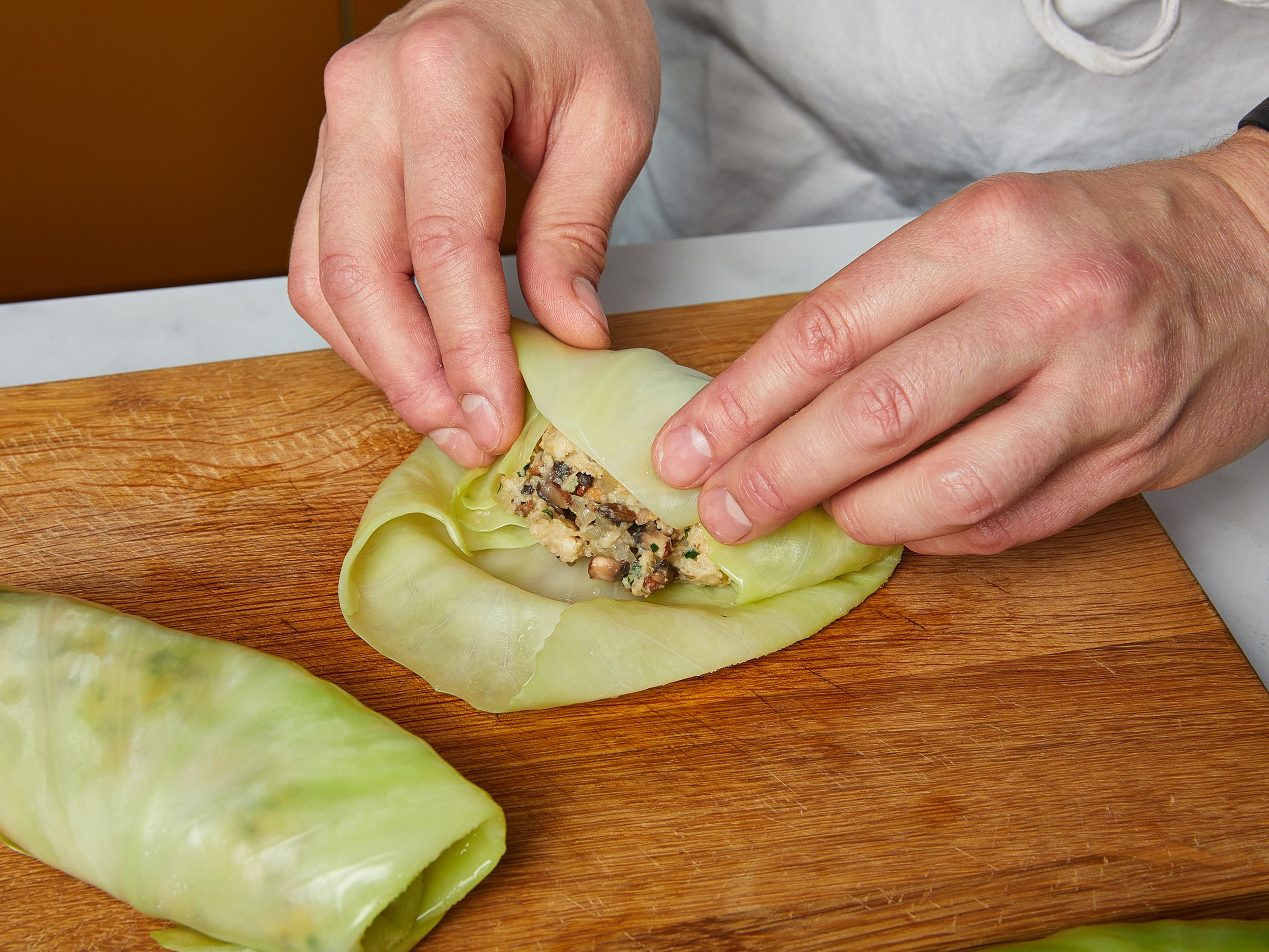 Add the bread cubes, half of the chopped parsley and the onion-milk mixture to a large bowl and gently knead into a sort of dumpling filling. Scoop approx. 2–3 tbsp of the filling onto the center of a cabbage leaf, roll it up and set aside. To do this, fold both sides over the filling and roll up from the stalk. Continue with the remaining cabbage leaves. If needed, seal cabbage rolls with a toothpick or some kitchen twine.