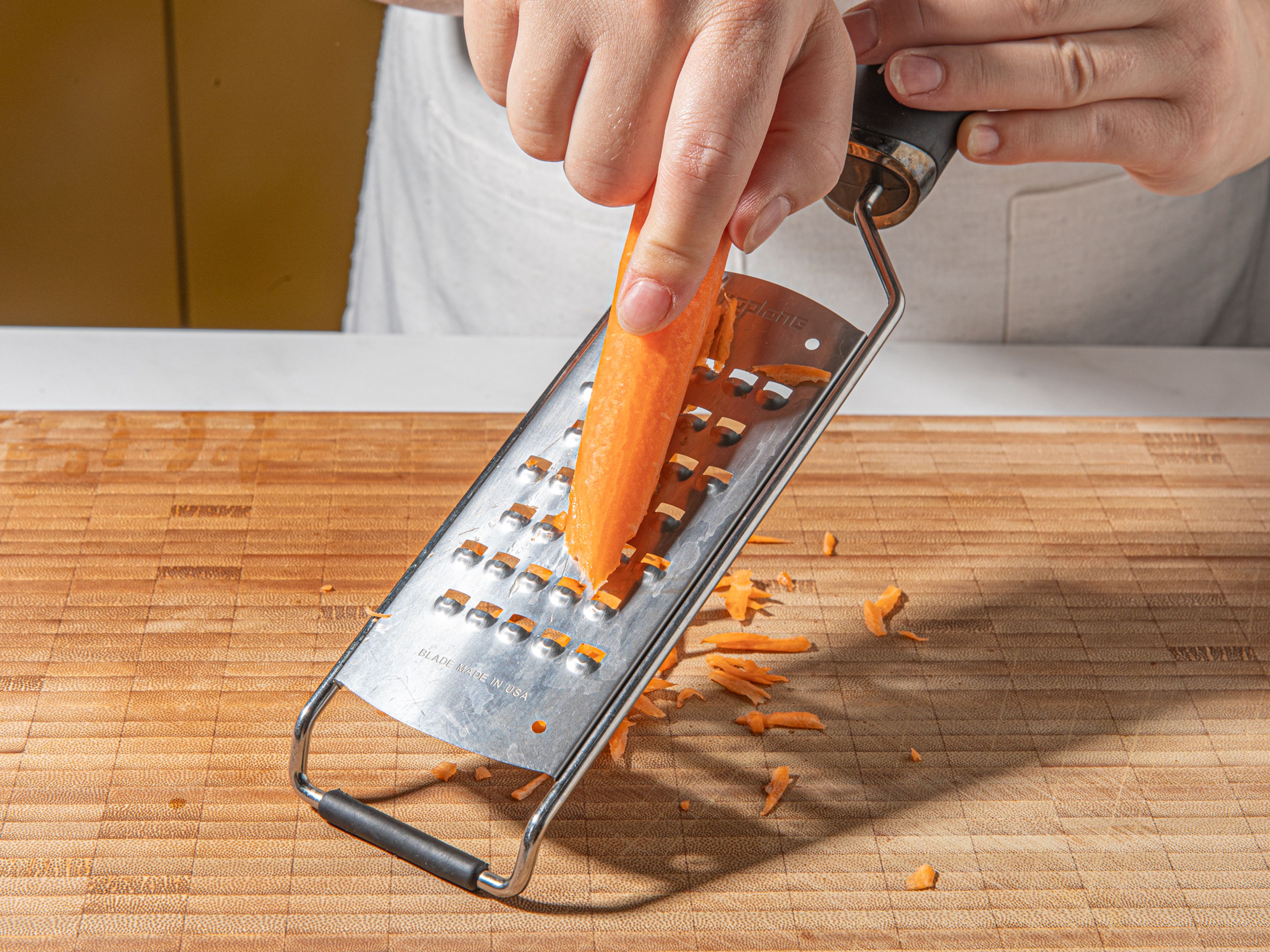 Das Ende des Salatkopfes abschneiden und die Blätter abtrennen. Die Karotte schälen, raspeln und in eine Schüssel geben. Mit Salz würzen und beiseite stellen. Gurken schräg in dünne Scheiben schneiden, sodass große, längliche Scheiben entstehen. Paprika vom Kerngehäuse befreien, entkernen und in dünne Scheiben schneiden. Tomate in dicke Scheiben schneiden. Minze und Petersilie abzupfen und grob hacken. Grüne Oliven ebenfalls hacken.