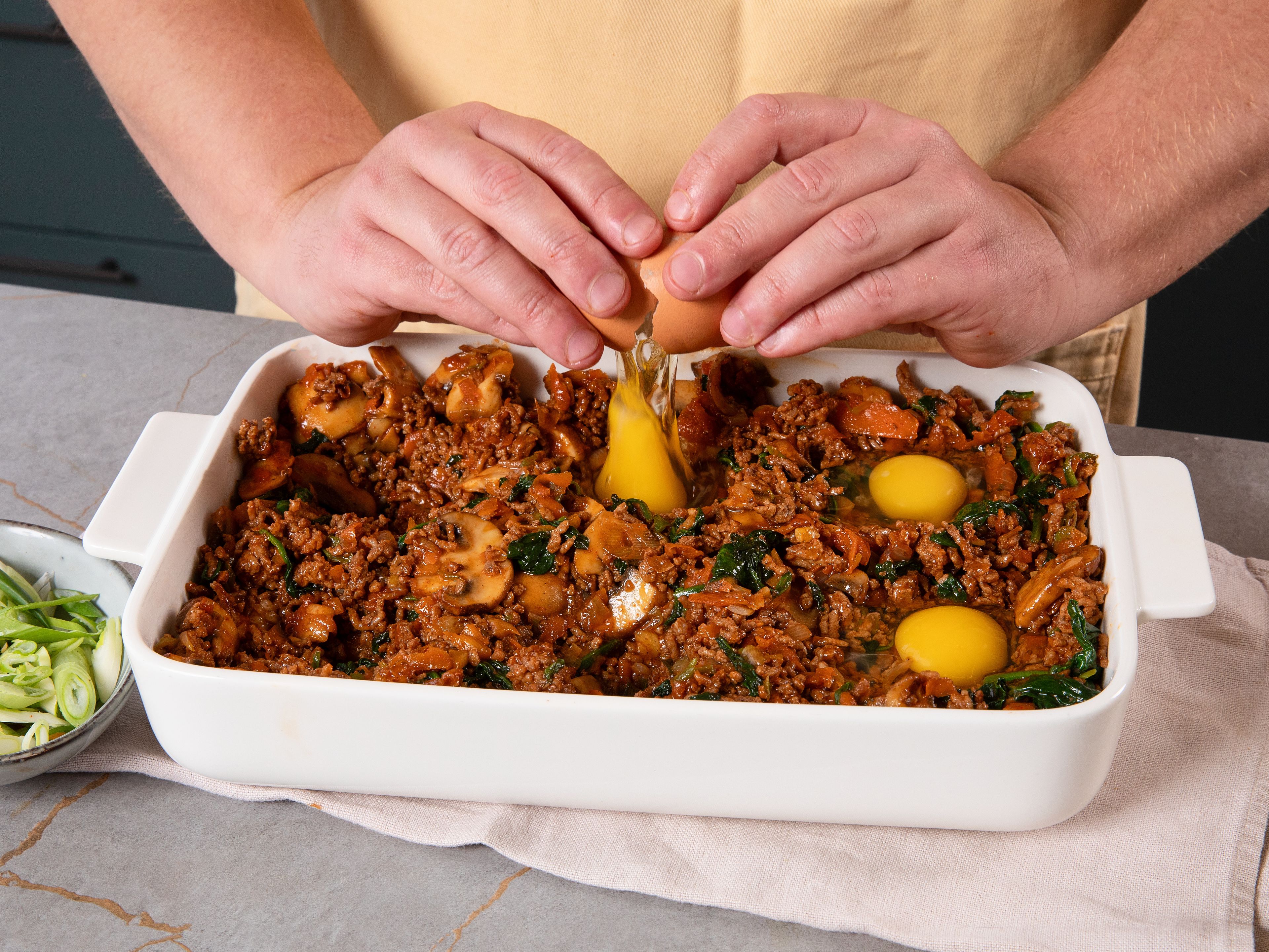Mit dem Rücken eines Löffels Vertiefungen für die Eier in die Mischung drücken. Jeweils ein Ei in jede Vertiefung schlagen. Dann die Auflaufform in den Ofen stellen und ca. 20 Min. backen, bis die Eier gestockt sind. Sobald sie nach deinem Geschmack fertig sind (ob hartgekocht oder noch etwas flüssig), aus dem Ofen nehmen. Mit Sriracha und Frühlingszwiebelringen garnieren und servieren.