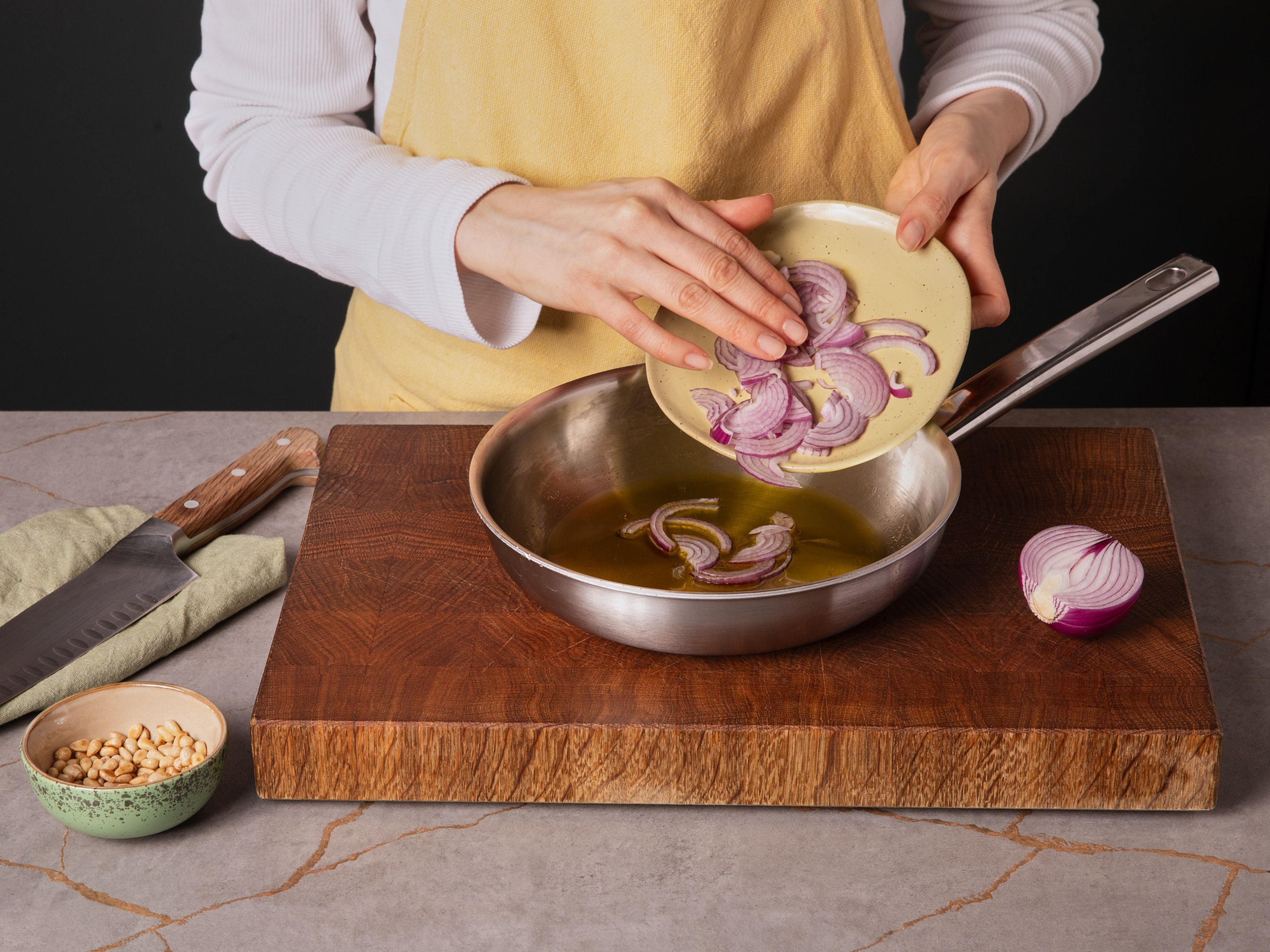 Pinienkerne in einer kleinen Pfanne goldbraun rösten, dann herausnehmen und beiseitestellen.
In derselben Pfanne Olivenöl erhitzen. Die Knoblauchzehe schälen, andrücken und kurz im Öl anbraten, um es zu aromatisieren. Knoblauch entfernen und für später aufbewahren.
Die Zwiebel in feine Halbmonde schneiden, in das noch warme, aromatisierte Öl geben und ziehen lassen.