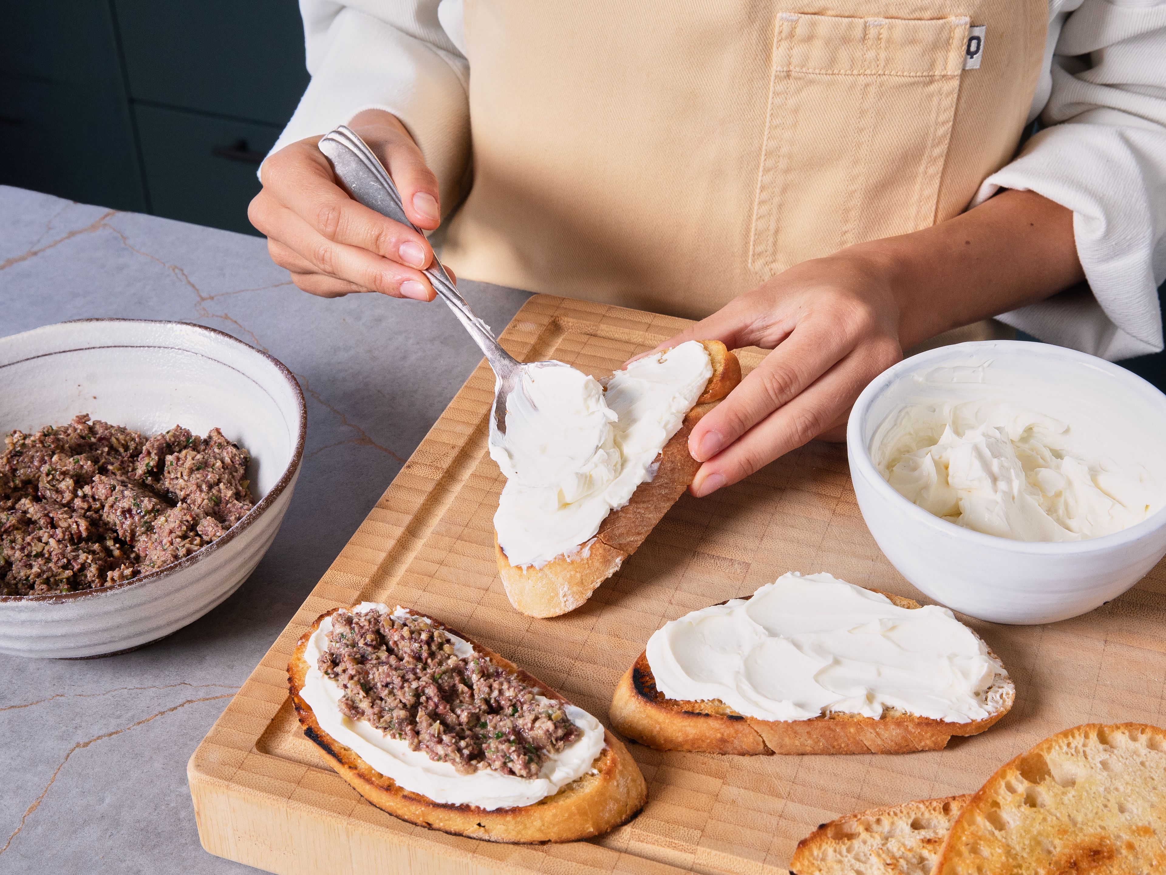 Frischkäse mit Salz und Pfeffer abschmecken. Brot, Crostini oder Cracker mit dem Frischkäse bestreichen und mit Oliven-Tapenade toppen.