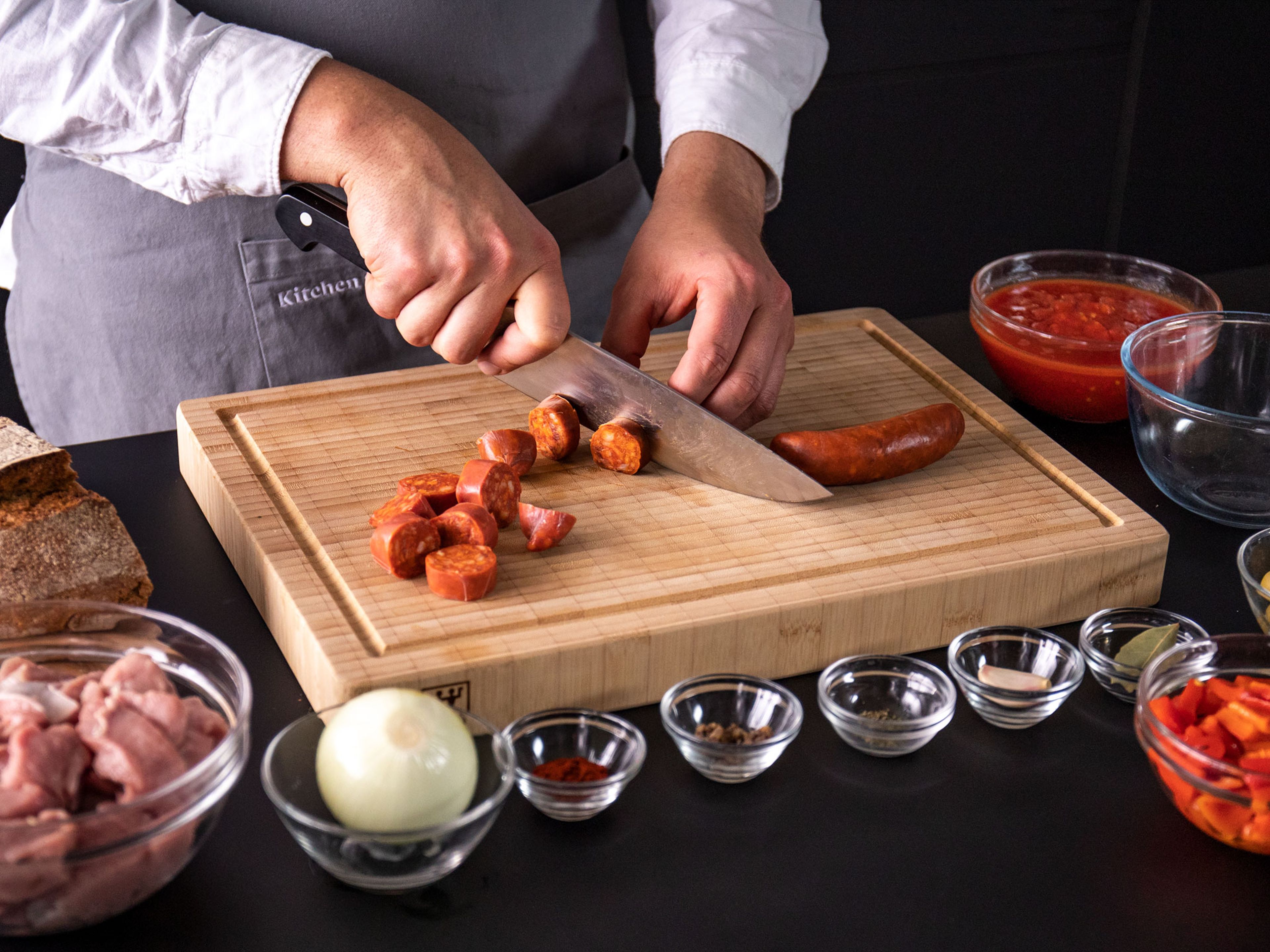 Zwiebel und Knoblauch schälen und fein hacken. Gegrillte Paprika aus dem Glas abtropfen lassen und klein schneiden. Chorizo in Scheiben schneiden und Sardellenfilets fein hacken.