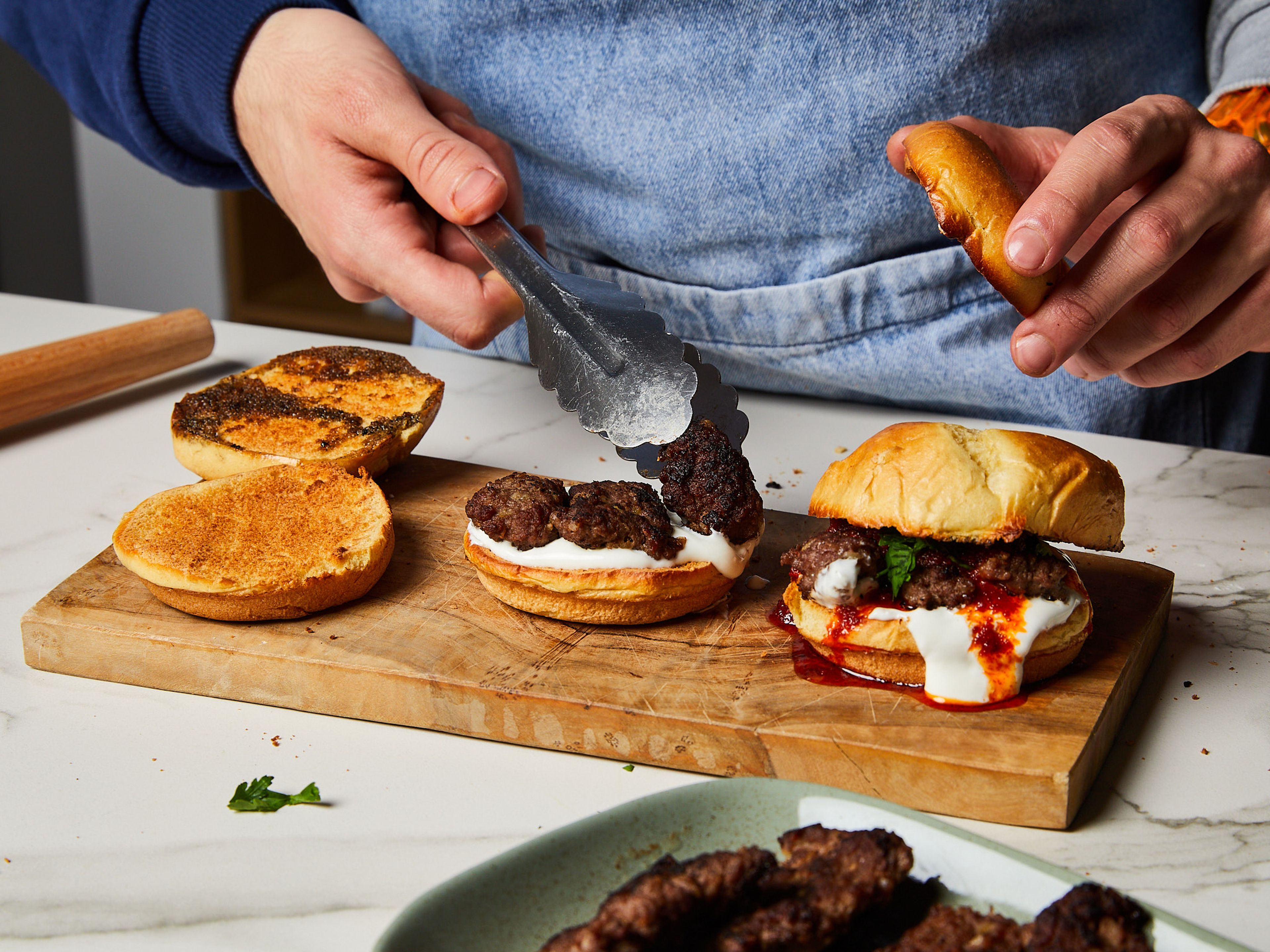 Nach dem Backen die Burger-Brötchen mit der Schnittseite in das restliche Öl der Pfanne drücken, falls noch etwas übrig ist. Dann schichten: Untere Brötchenhälfte, Joghurtsoße, 2–3 Köfte-Stücke, eine Prise Salz, buttrige Tomatensauce, gehackte Petersilie und zum Schluss die obere Brötchenhälfte. Leicht andrücken und mit den restlichen Brötchen wiederholen und sofort servieren!