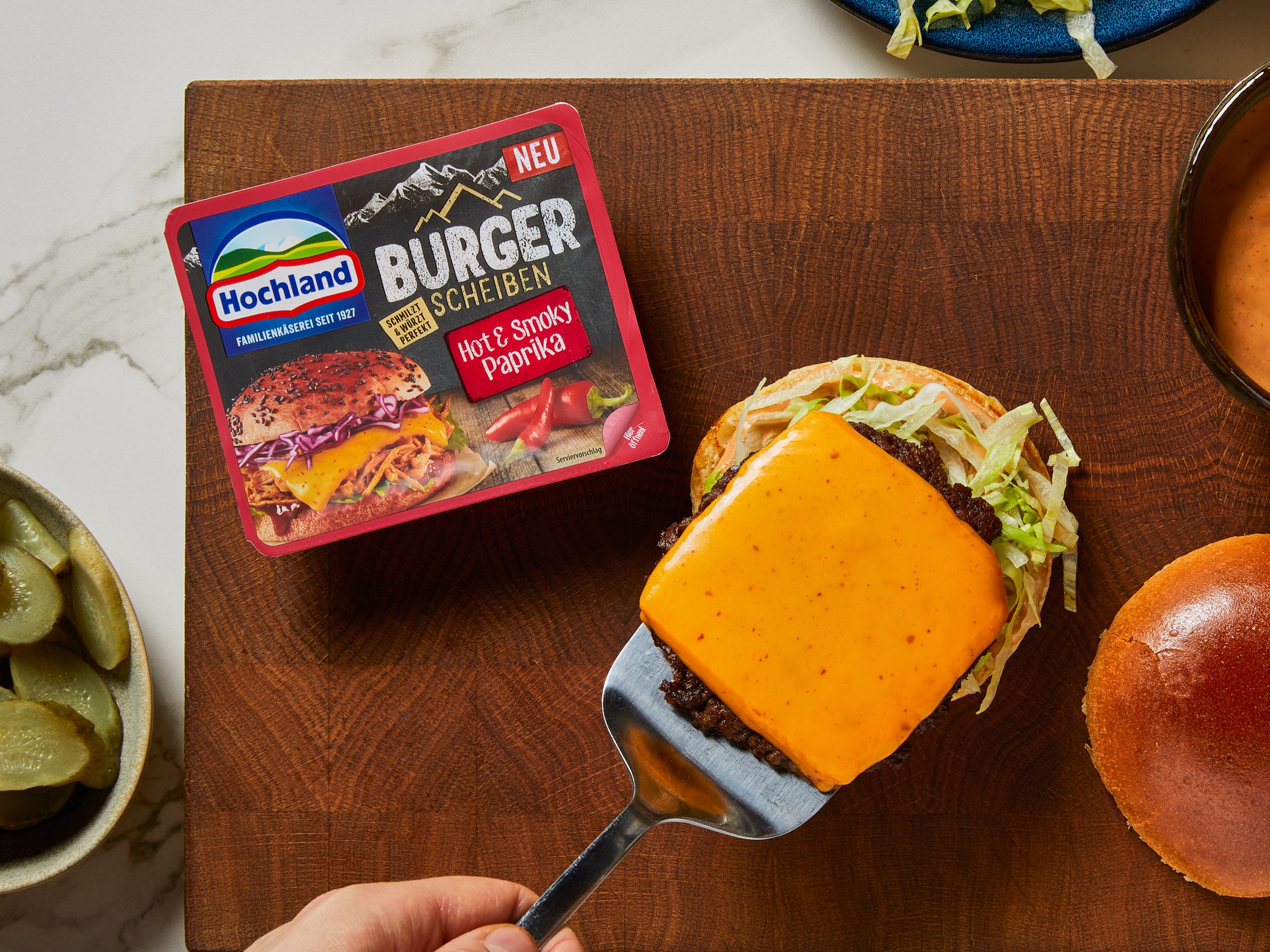 Die Smash Burger-Patties wenden, mit Salz und Pfeffer würzen und mit jeweils einer Hochland Burger Scheibe Hot & Smoky Paprika belegen. Weitere 1–2 Min. garen. In der Zwischenzeit die Brötchen auf beiden Seiten mit der Soße bestreichen und die untere Hälfte mit dem Salat belegen. Dann die Patties, Zwiebeln, Gurkenscheiben und den oberen Teil des Brötchens darauf legen. Sofort servieren!