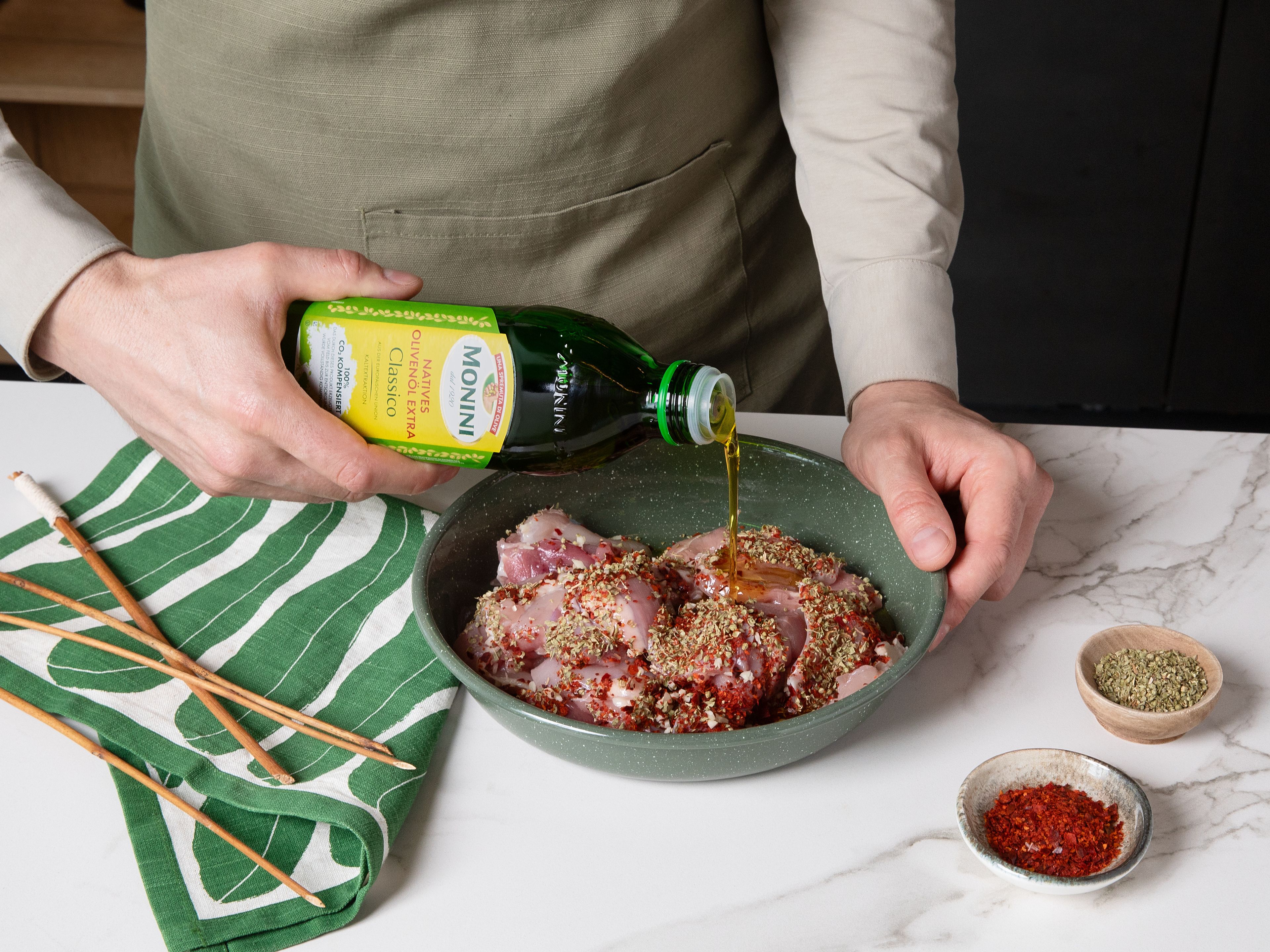 Knoblauch fein hacken.  Das Hähnchen in mundgerechte Stücke schneiden und in eine Schüssel geben. Mit 4 EL Monini Classico Olivenöl, Knoblauch, Salz, Pfeffer, Oregano und Chiliflocken vermengen. Die Fleischstücke auf Spieße stecken und für mindestens 30 Minuten ziehen lassen.