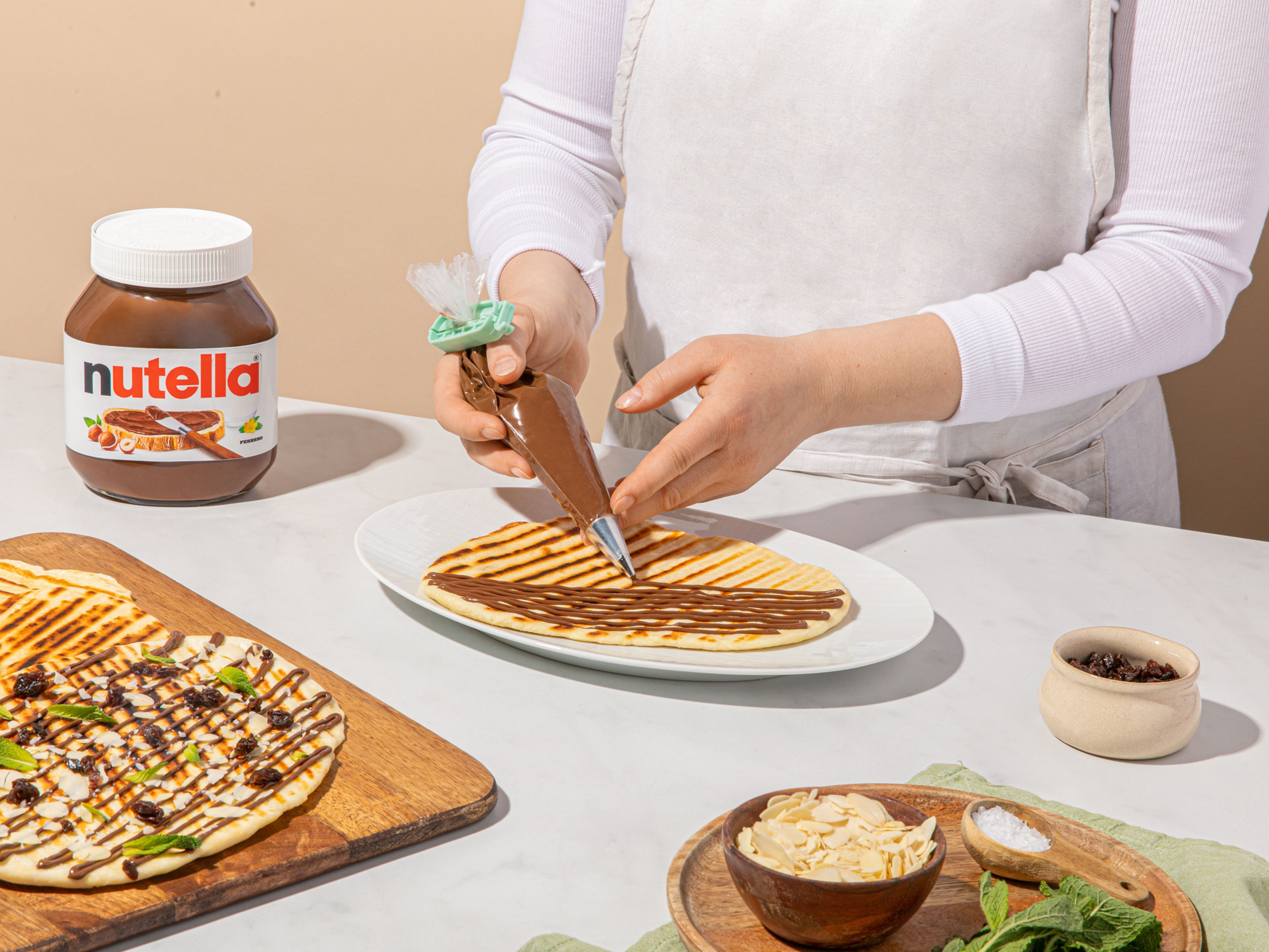 nutella® in einen Spritzbeutel füllen und vorsichtig in Linien über die Naan verteilen. Mit Rosinen, Mandeln, Minze und etwas Meersalz garnieren.