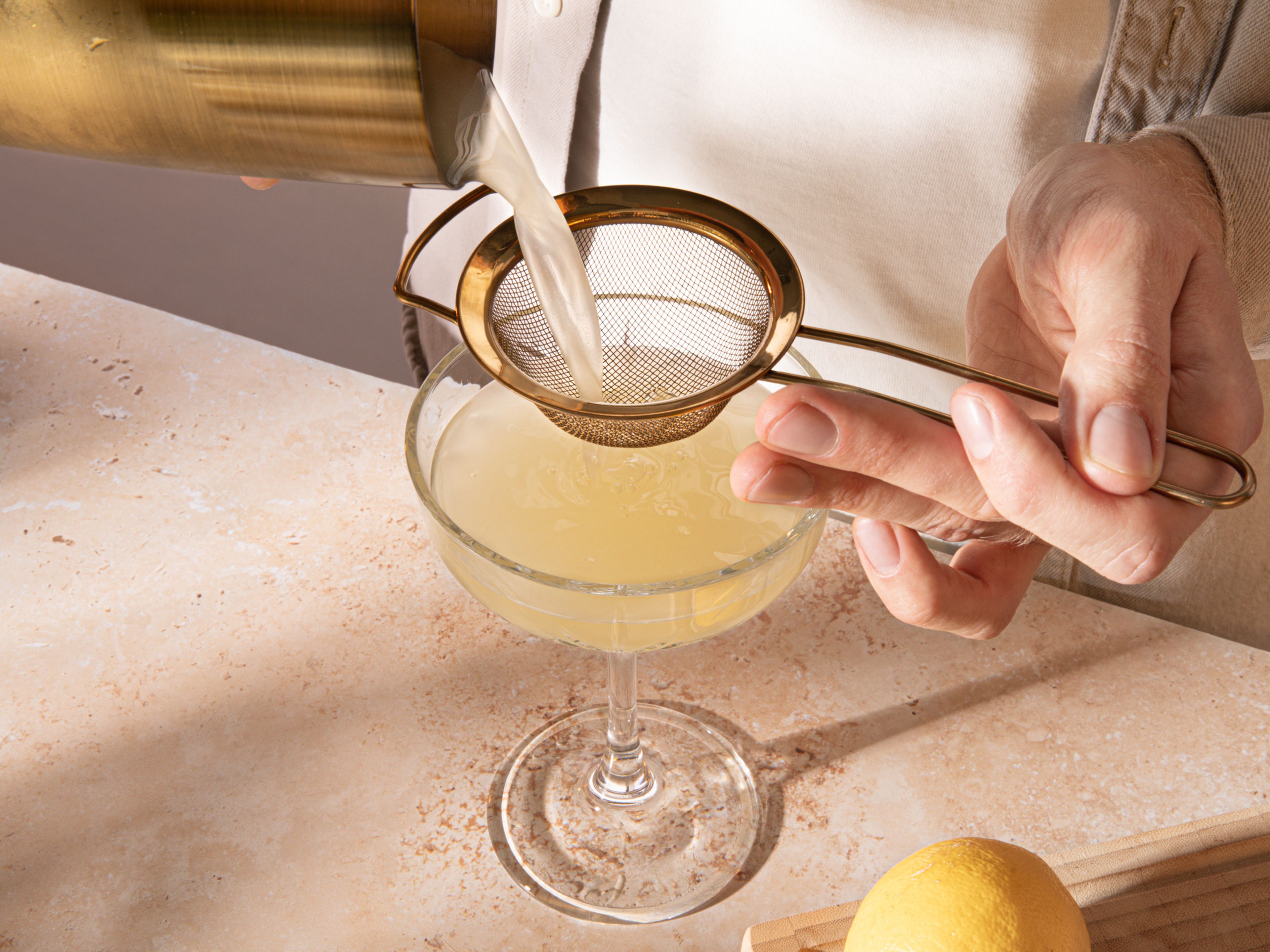 Durch ein Barsieb und ein zusätzliches Sieb in ein Glas abseihen, um kleine Eisstücke abzufangen. Mit Sekt aufgießen, Zitronenschale auf einen Cocktailspieß spießen und den Drink damit dekorieren.