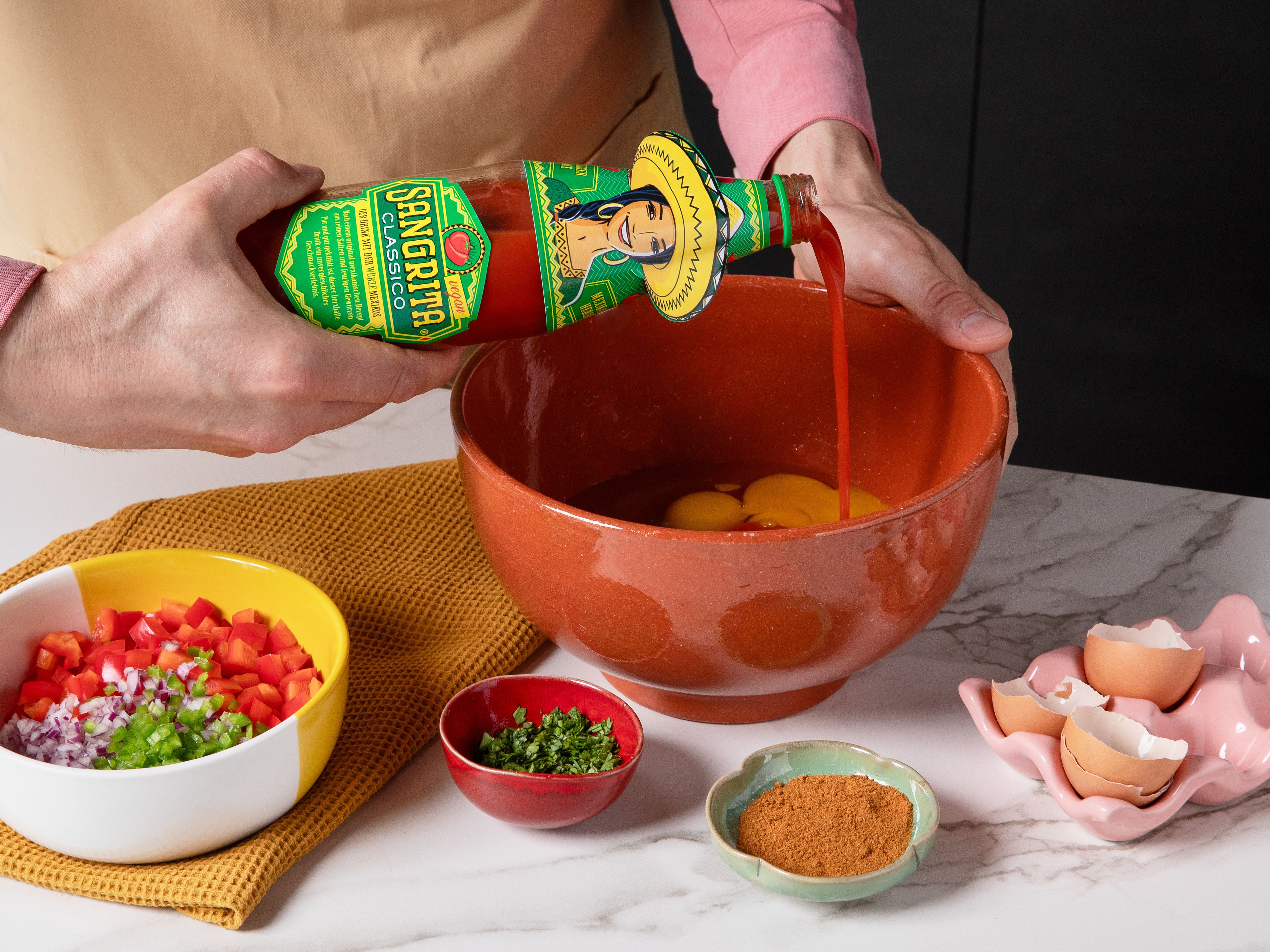 In einer großen Schüssel die Eier mit Sangrita verquirlen, dann Paprika, Zwiebel, Jalapeño und zwei Drittel des Korianders untermischen. Mit Salz, Pfeffer und nach Belieben etwas gemahlenem Kreuzkümmel würzen. Eine ofenfeste Form oder ein kleines Backblech mit Backpapier auslegen, die Eiermasse hineingießen und glatt streichen. Den geriebenen Cheddar gleichmäßig darüberstreuen und im Ofen ca. 18–20 Min. backen, bis die Eier gestockt und goldgelb sind.