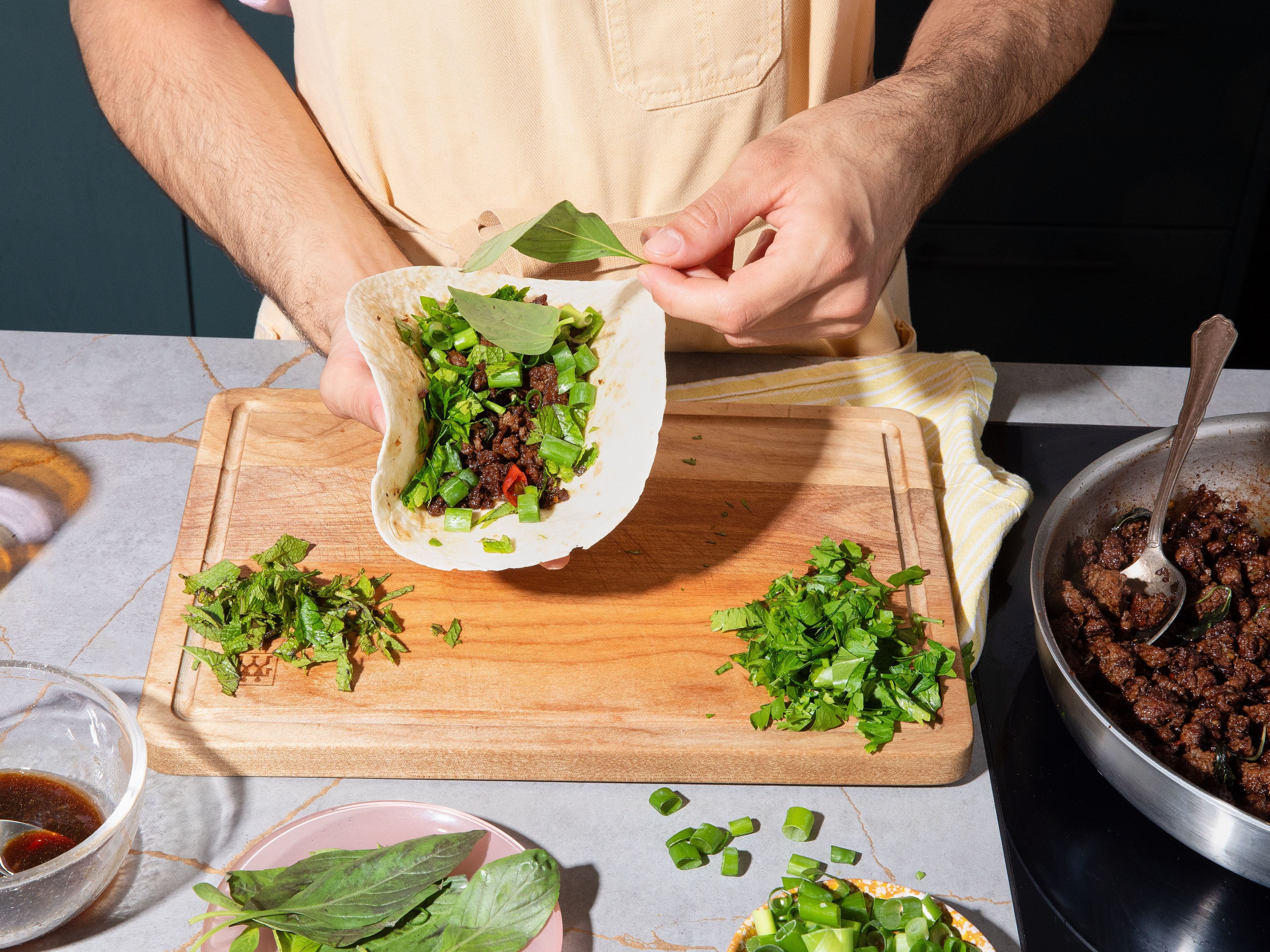 Once the beef is cooked, remove from heat. Place the tortillas on top of the beef mixture briefly to warm them up and let them absorb some of the flavorful fat. Fill each tortilla with the beef mixture, then top with mint, cilantro, green parts of the scallions, and fresh Thai basil. Roll up and serve immediately with the dipping sauce.