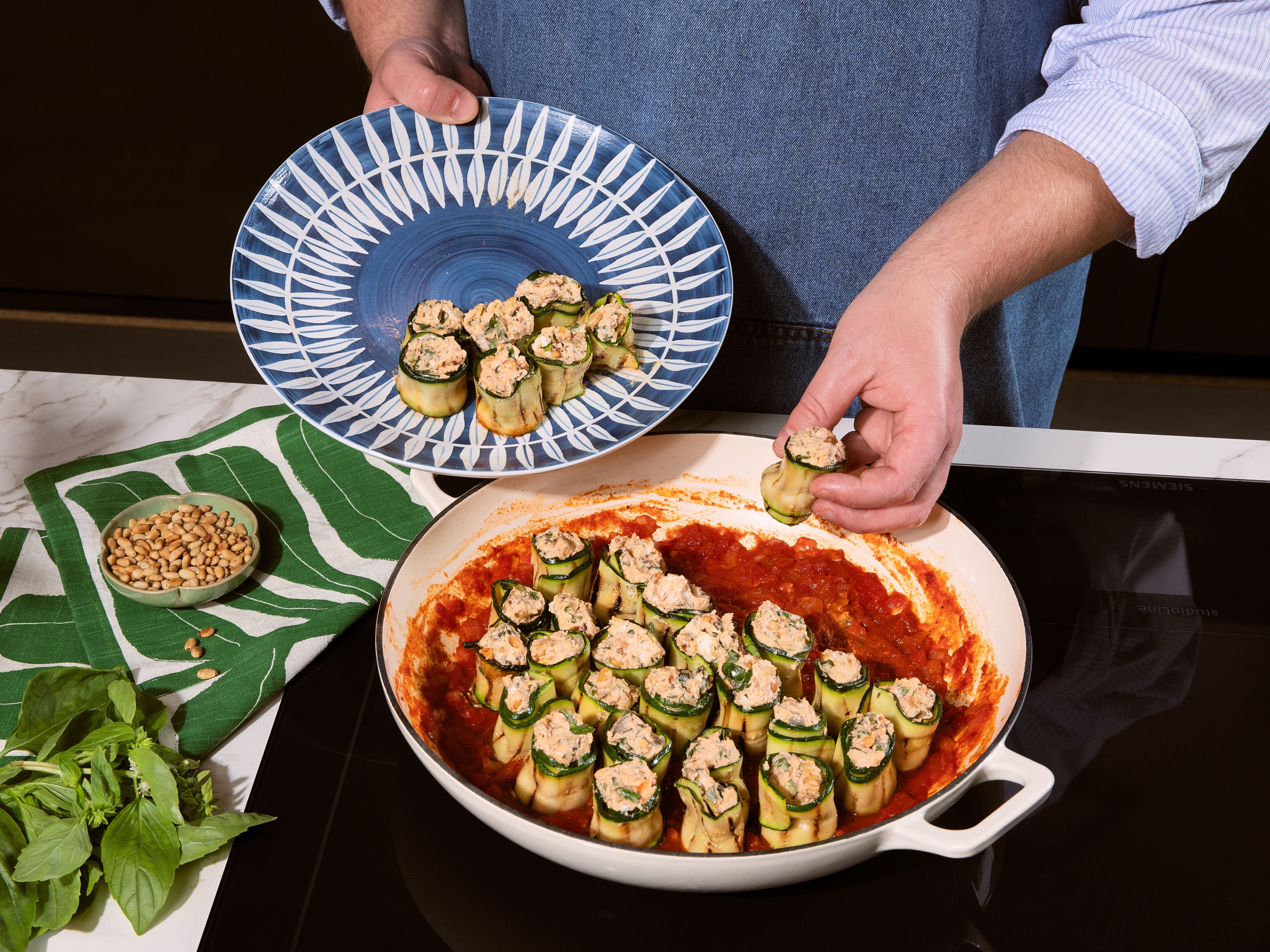 Zwiebel und Knoblauch fein hacken. In einer ofenfesten Pfanne etwas Olivenöl erhitzen und die Zwiebel darin ca. 3 Min. glasig dünsten. Knoblauch hinzufügen und ca. 1 Min. mitbraten. Gehackte Tomaten dazugeben und ca. 5 Min. köcheln lassen. Mit Salz, Pfeffer und einer Prise Zucker abschmecken. Zucchiniröllchen dicht an dicht hochkant in die Tomatensauce setzen, bis die Pfanne gefüllt ist. Mit etwas Parmesan bestreuen und im vorgeheizten Ofen ca. 15 Min. backen, bis der Käse geschmolzen ist. Mit den restlichen Pinienkernen und dem übrigen Basilikum garnieren und servieren.