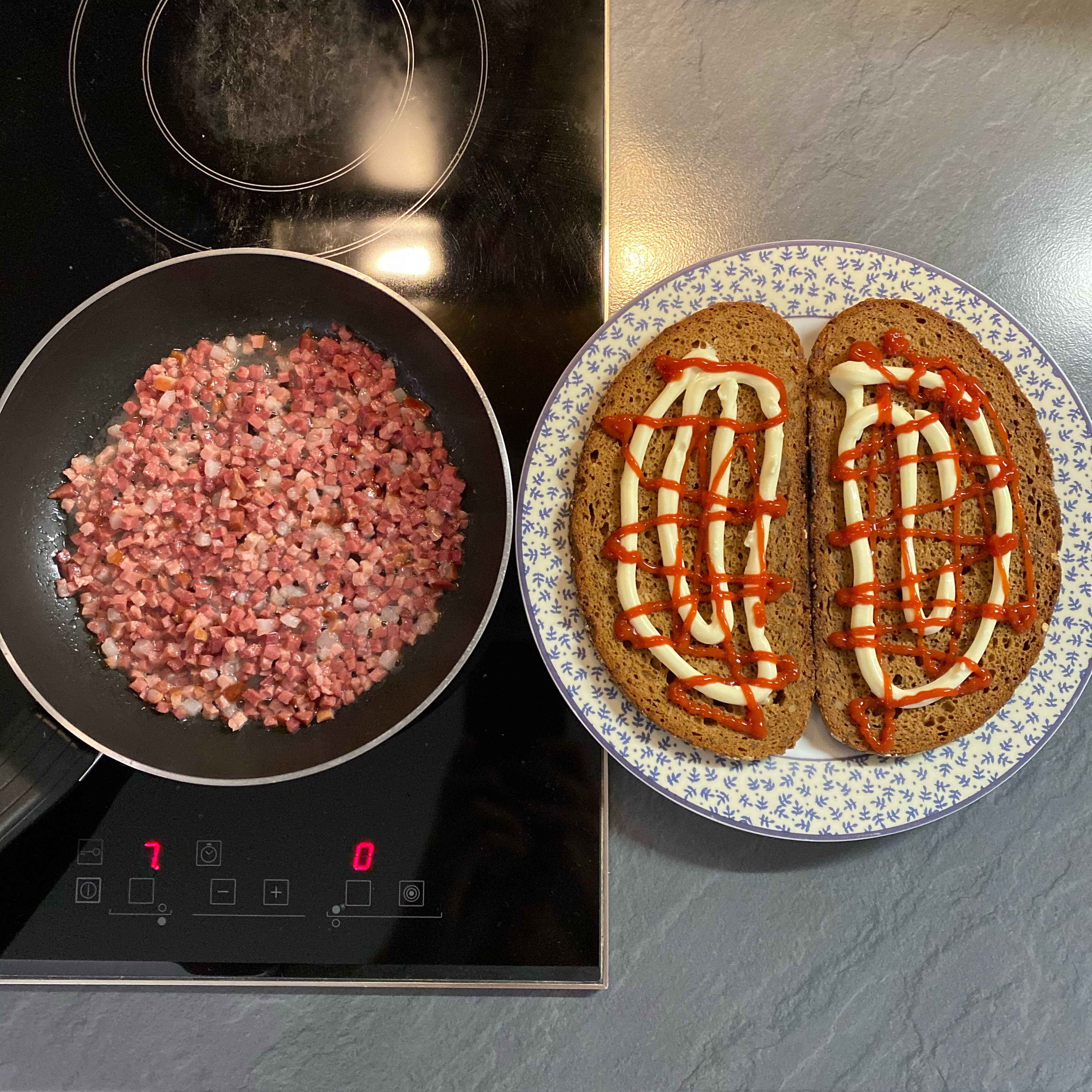 Den Bacon (Streifen oder Würfel) in ein wenig Öl anbraten. Währenddessen das Vollkornbrot auf einen Teller legen und teilweise mit Ketchup und Mayonnaise bedecken.
