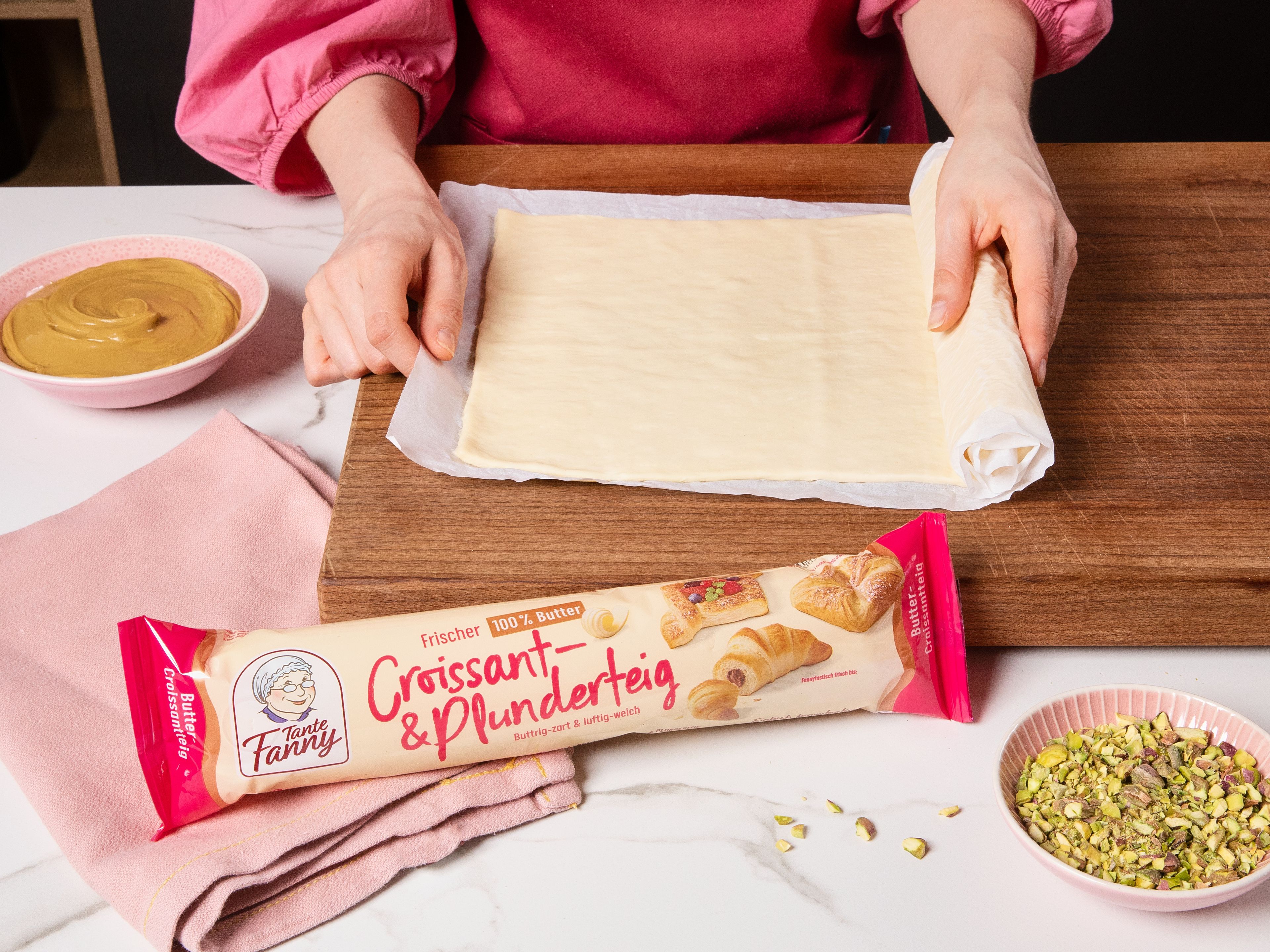 Backofen auf 180 °C Ober-/Unterhitze vorheizen. Den Tante Fanny Frischen Butter-Croissant- & Plunderteig ausrollen und glatt streichen. Den Großteil der Pistaziencreme gleichmäßig auf dem Teig verteilen. Die Pistazien grob hacken. Einen Teil der gehackten Pistazien zur Deko beiseite legen und den Rest gleichmäßig über die Creme streuen. Einen weiteren Teig vorsichtig ausrollen, darauflegen und leicht andrücken.