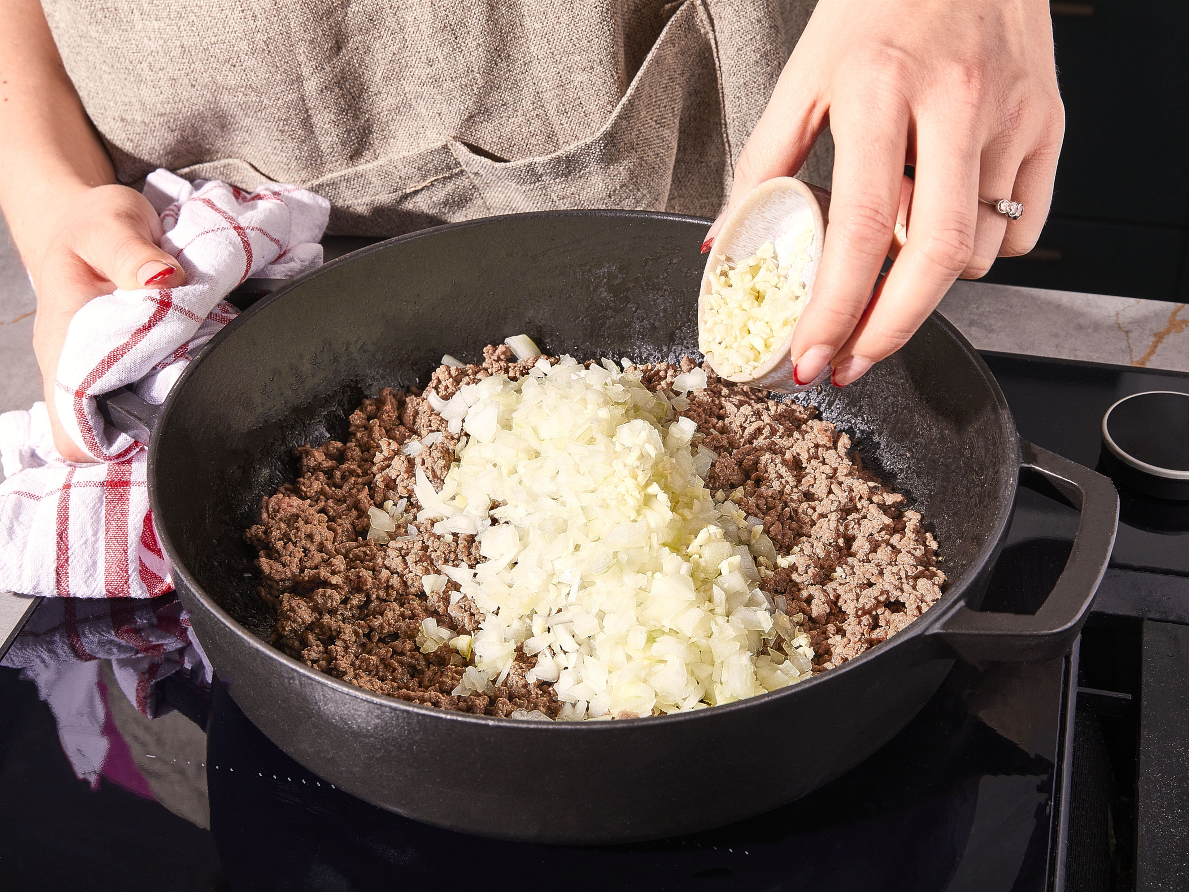 In der Zwischenzeit das Olivenöl bei mittlerer Hitze in einer großen Pfanne (mit Deckel) erhitzen. Das Rindfleisch hinzufügen und großzügig mit Salz und Pfeffer würzen. Das Rindfleisch anbraten und dabei mit einem Pfannenwender zerkleinern, damit es nicht kleben bleibt. Sobald das Rindfleisch anfängt, braun zu werden (ca. 5 Min.), Knoblauch und Zwiebeln hinzugeben. Ca. 3 Minuten weiterbraten, bis die Zwiebeln glasig werden.