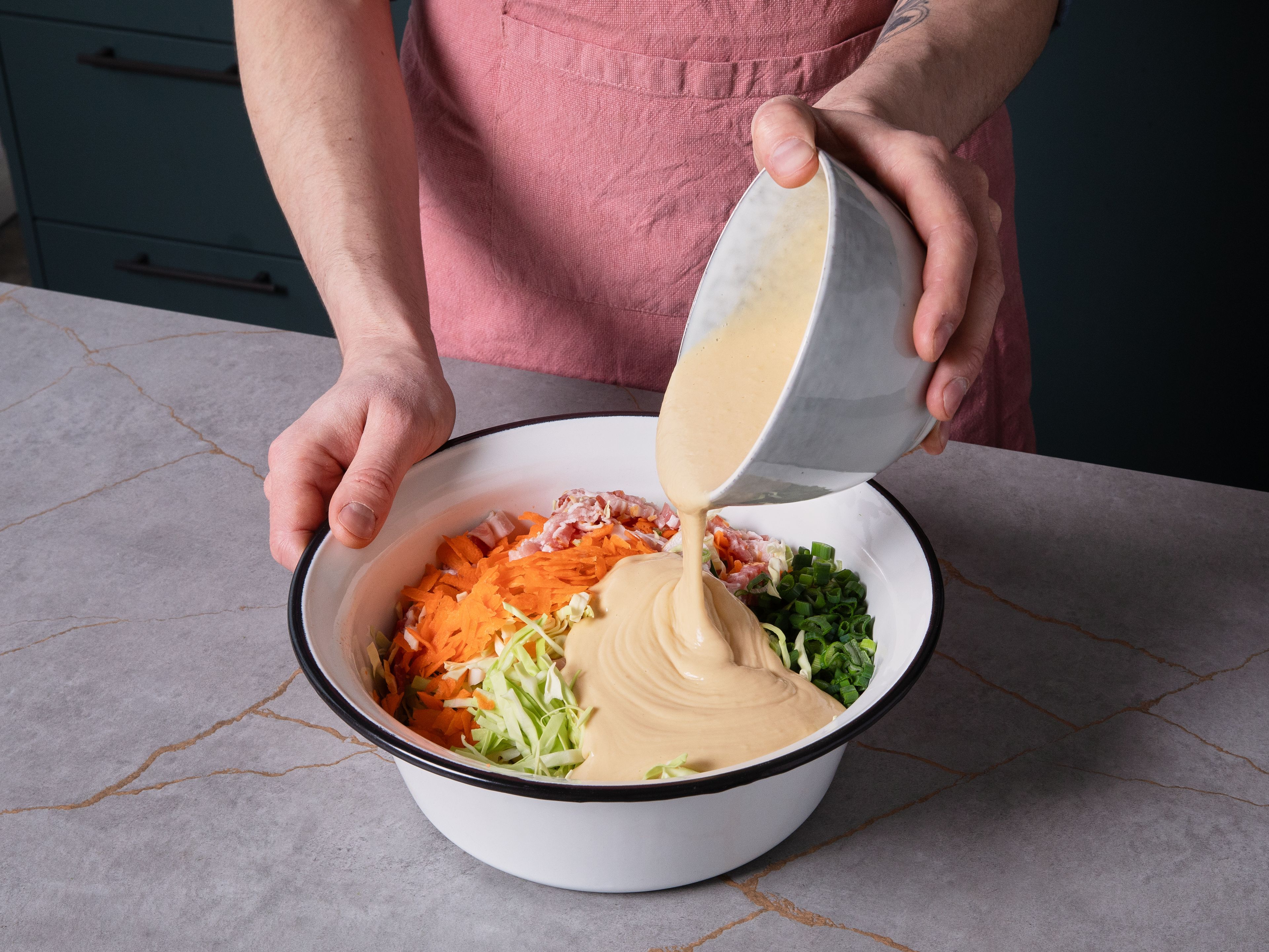 Den geschnittenen Kohl, die geriebenen Karotten und die Frühlingszwiebeln unter den Teig heben, sodass alles gut vermischt ist. Anschließend die Speckstücke hinzufügen und gleichmäßig vermengen.