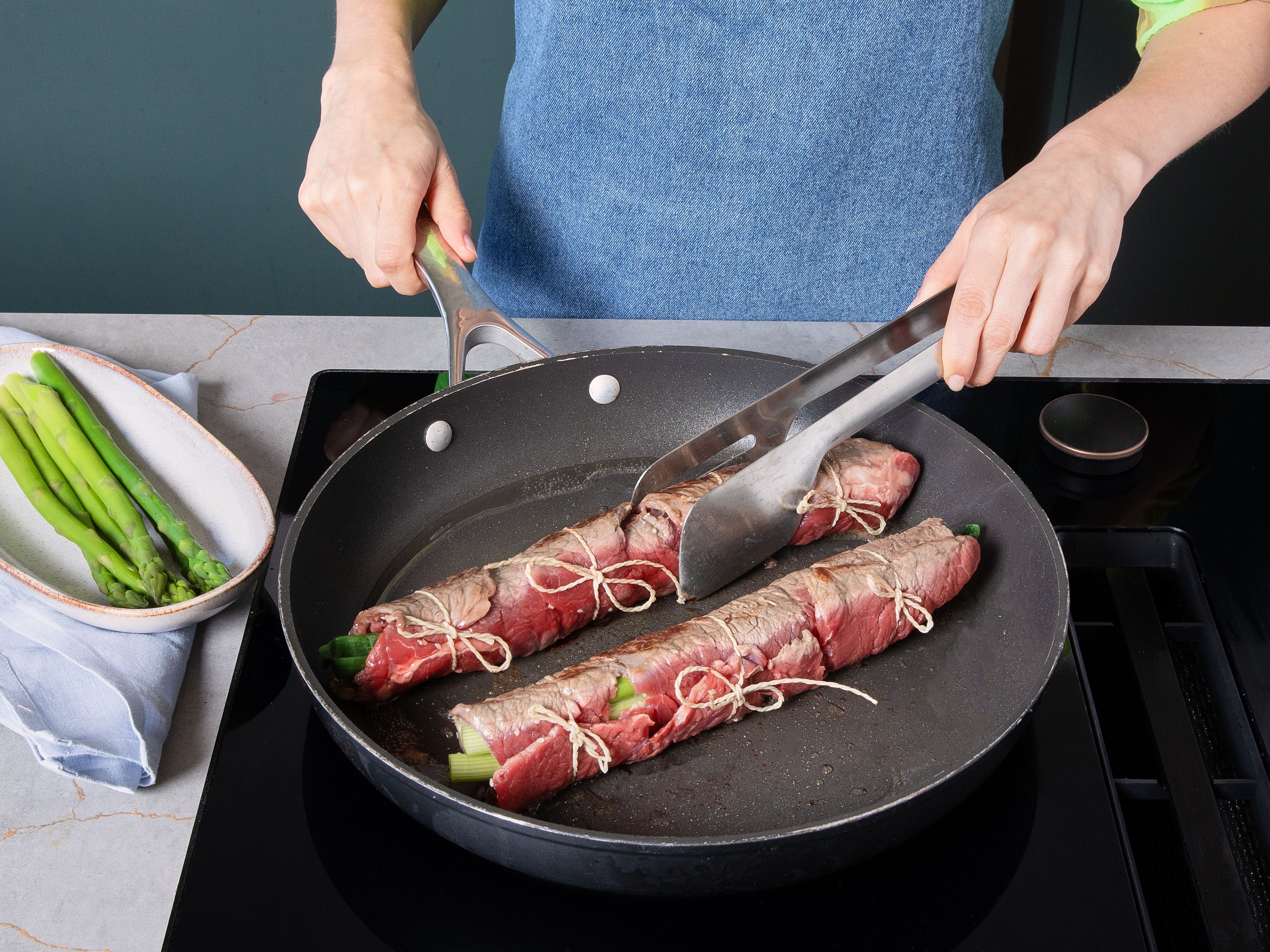 Place a few asparagus spears and spring onions on lower side of steaks and roll up tightly using film. Proceed same way with remaining ingredients, reserving some asparagus for serving. If necessary, secure rolls with kitchen twine. Heat vegetable oil in a large pan and sear rolls all around. Then reduce heat.